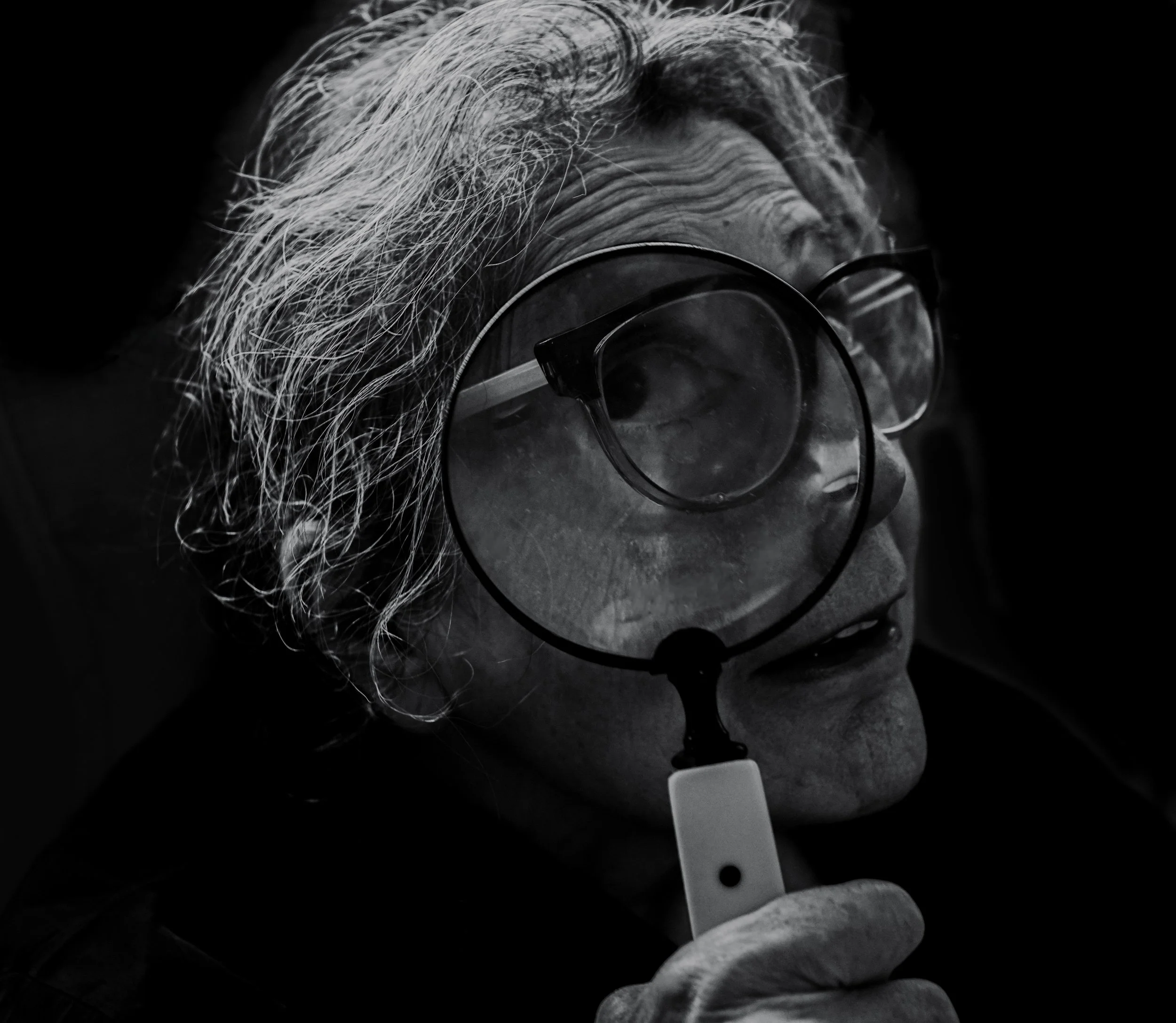 Close-up black and white photo of an elderly woman with glasses, looking with a magnifying glass held close to her eye.