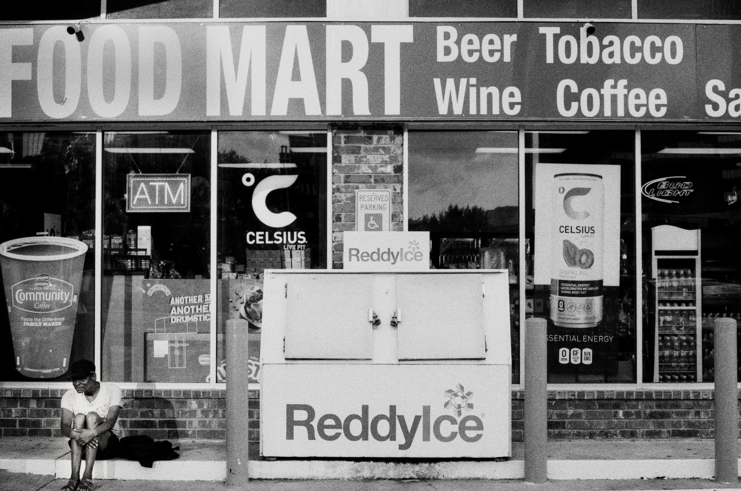 A woman sitting alone on a ledge in front of a convenience store, with signs for food, drinks, and cigarettes displayed on the store window, and advertisements for Celsius energy drinks.