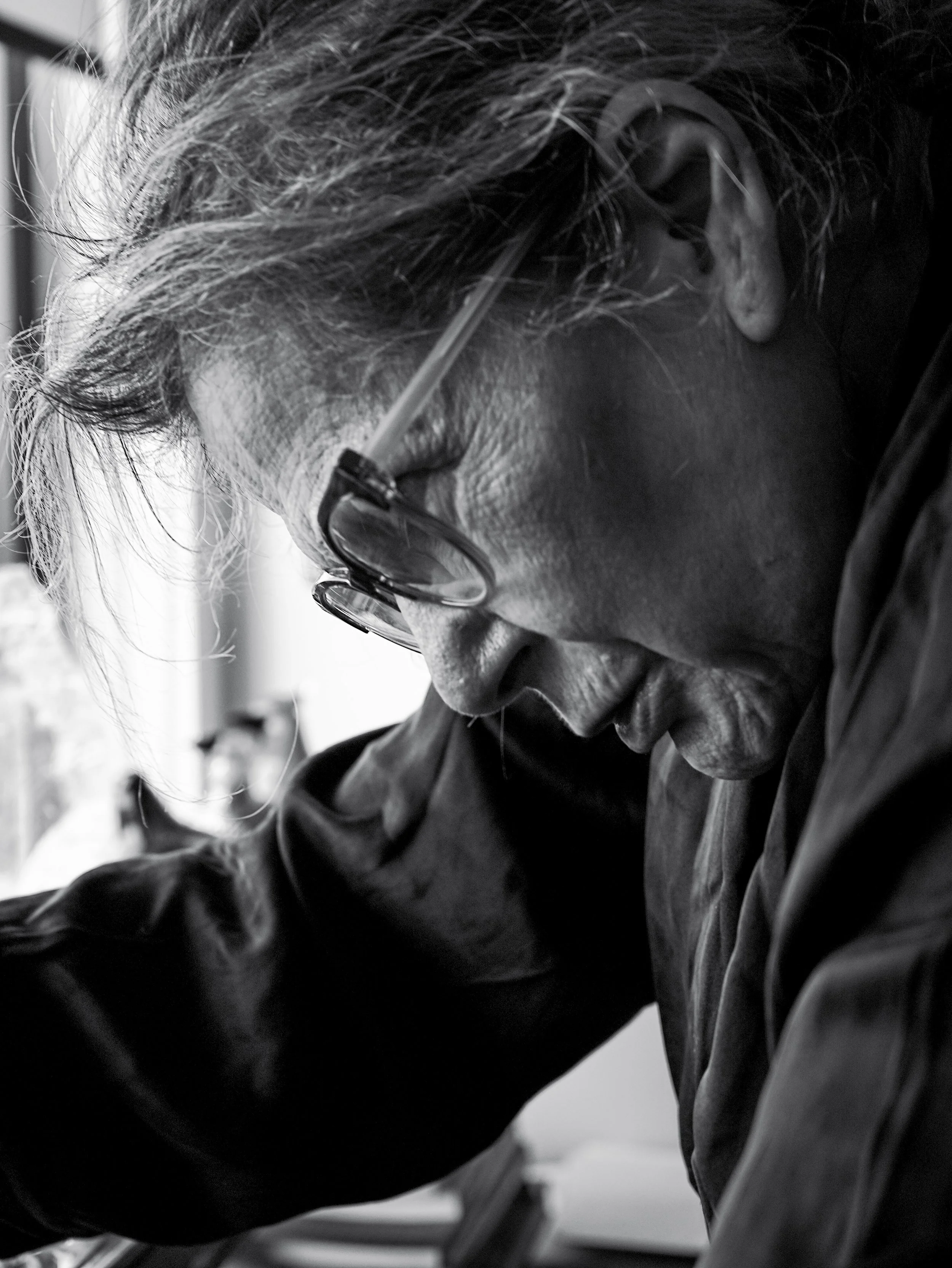 Close-up of an elderly man with glasses, looking down and smiling, with a window and blurred background.