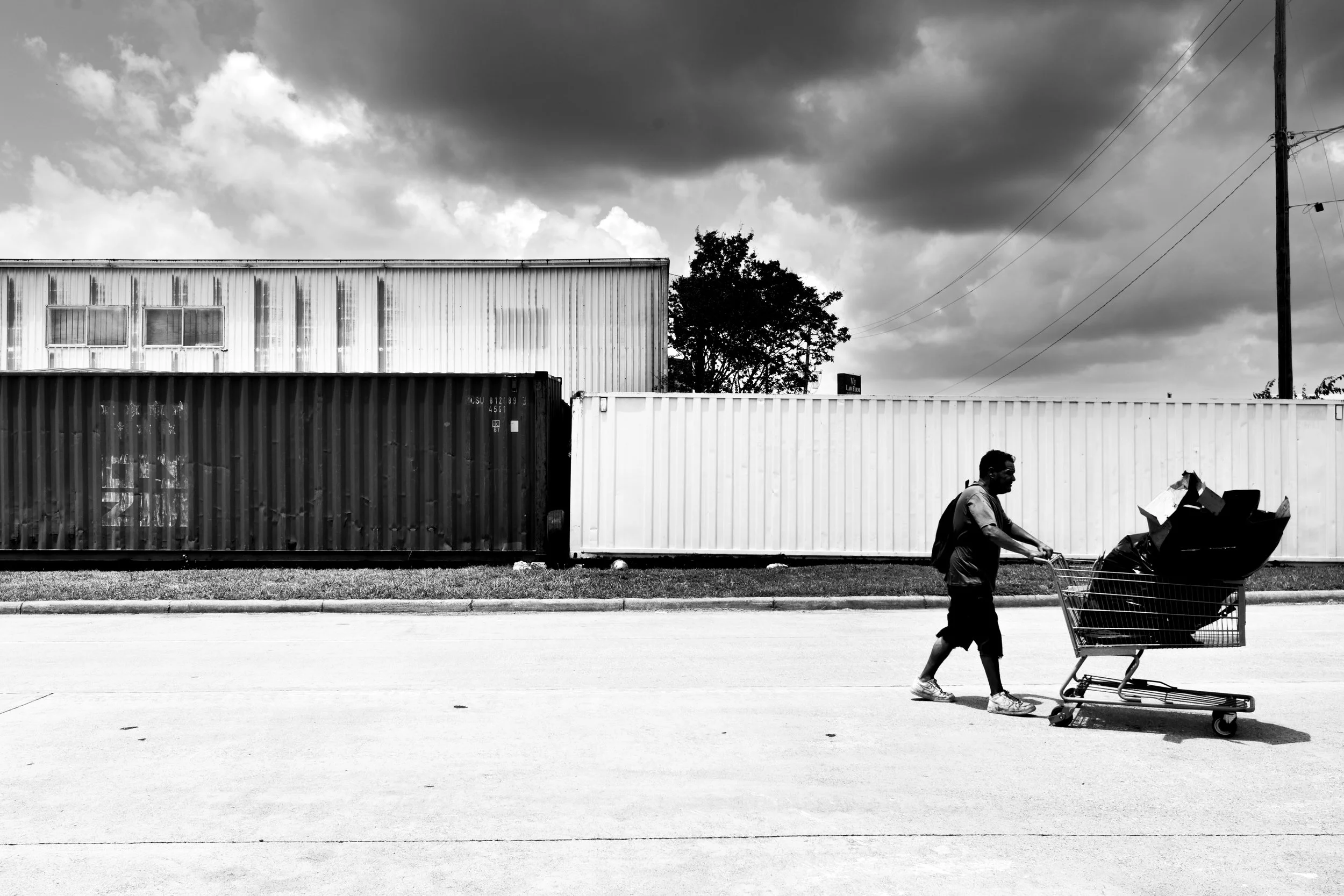 A man pushing a shopping cart filled with bags and boxes along a street in front of a fence made of shipping containers. The sky is cloudy, and there are some trees and a utility pole in the background.