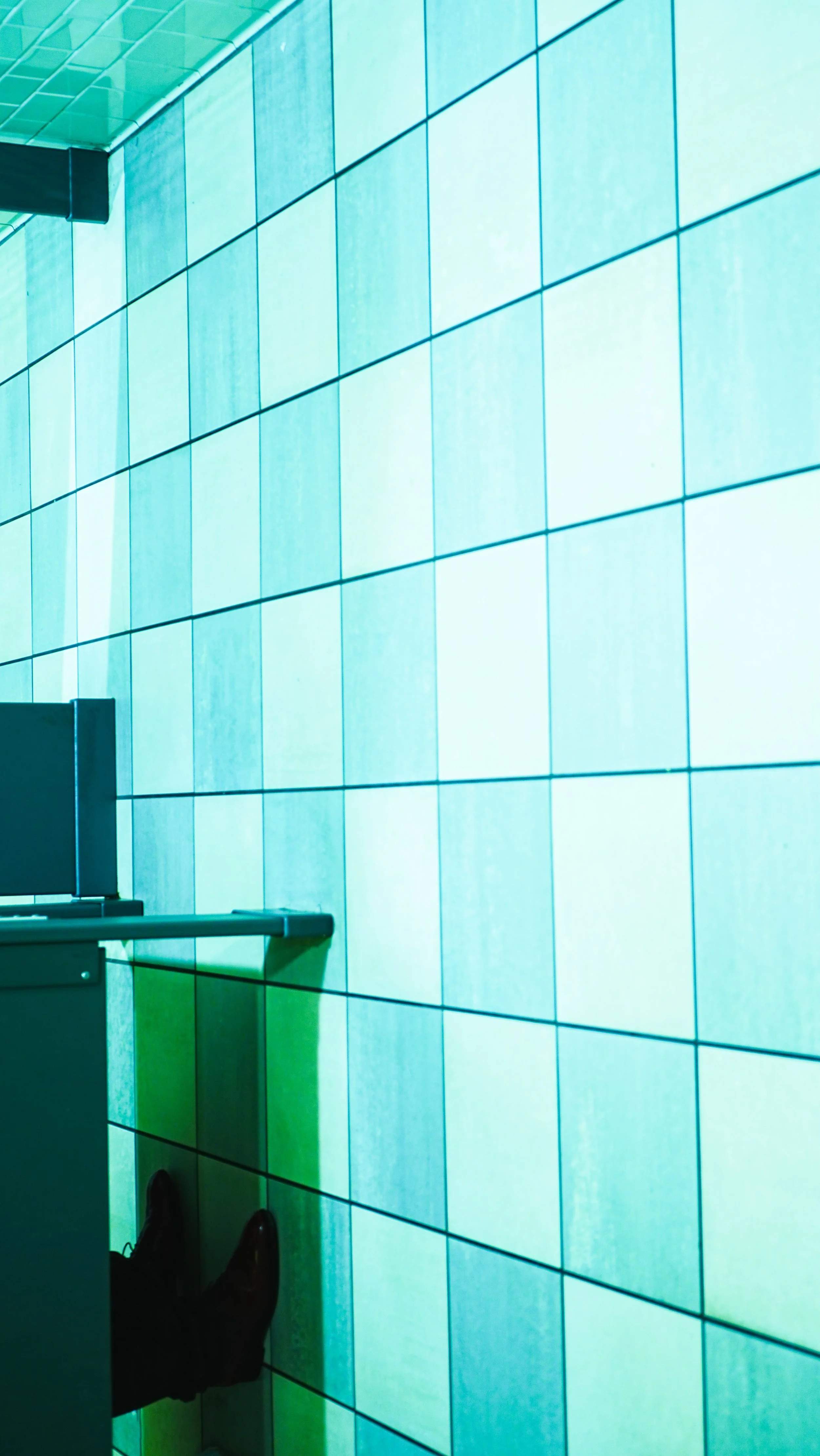 A modern interior with a large illuminated turquoise tiled wall, black monitor, white desk, and a person's legs and shoes visible at the bottom left.