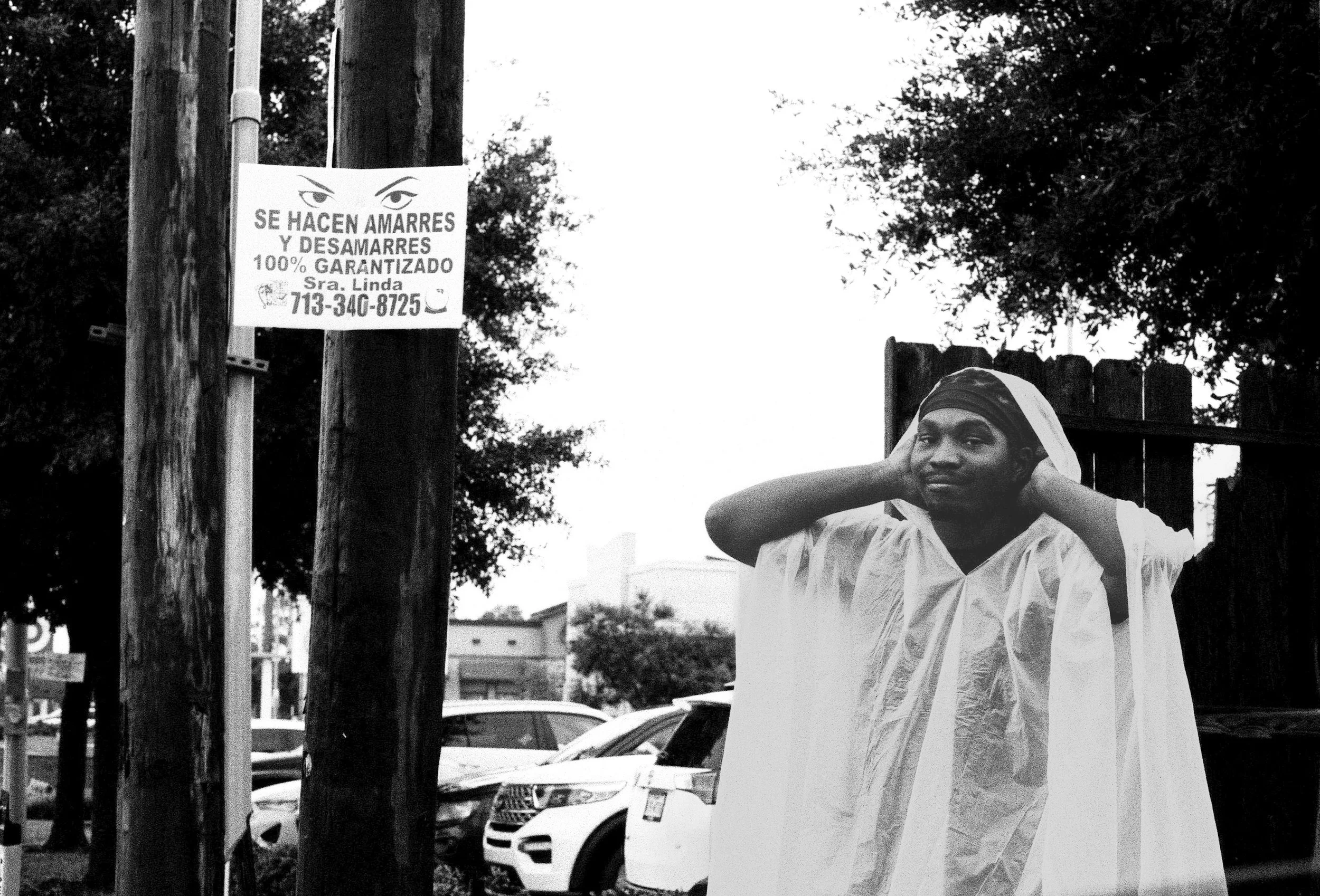 A woman wearing a white ceremonial gown, possibly a graduation or religious robe, standing outdoors near a wooden fence and car. She is holding her head with both hands and looking slightly to the side. There's a sign on a utility pole with text in S