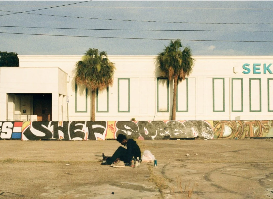 A person sitting on the ground in an empty lot, surrounded by graffiti art on a low wall, with two palm trees and a building in the background.
