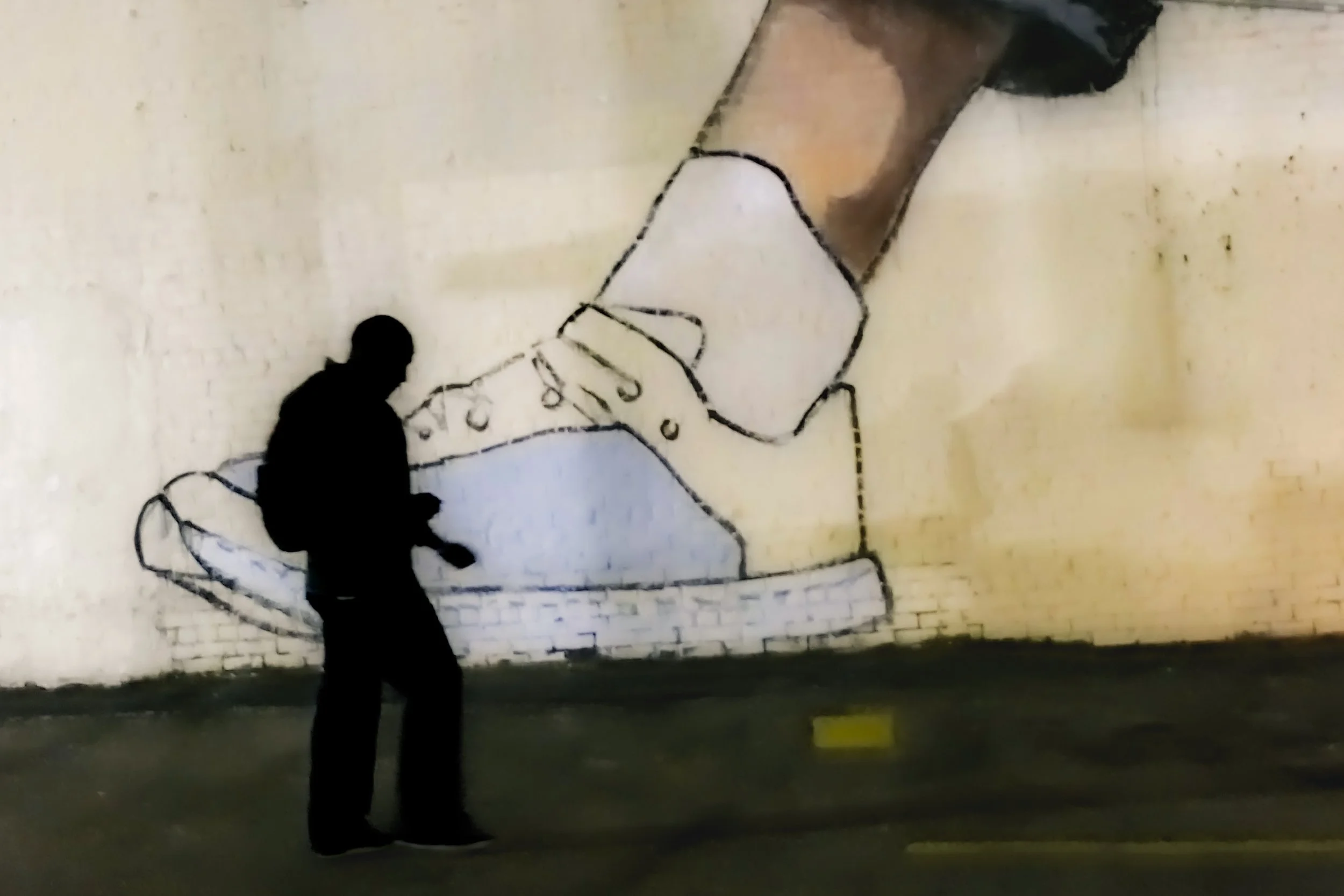 Silhouette of a person walking past a wall with street art of a giant sneaker shoe outlined in black and filled with beige, white, and light blue colors.
