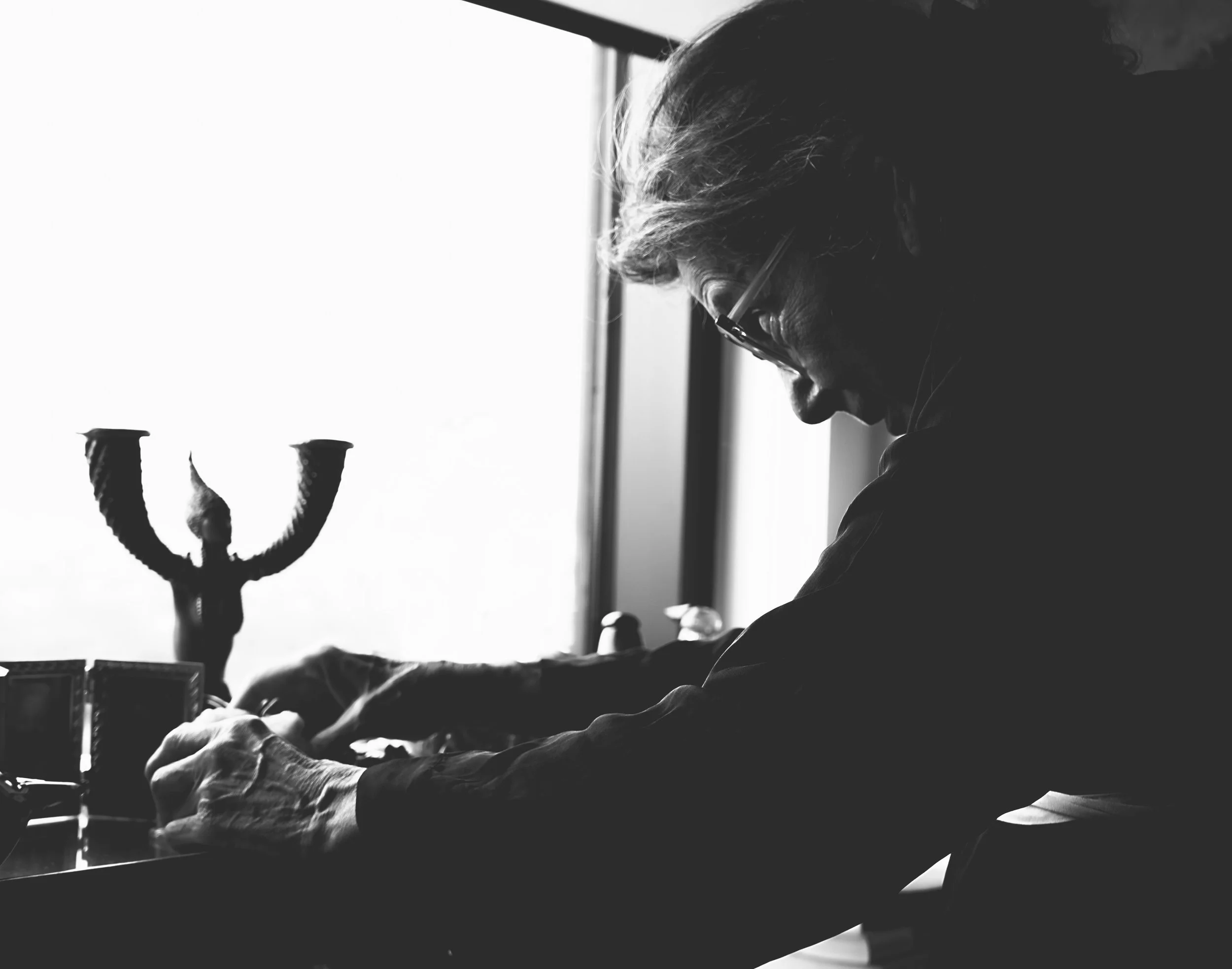 A black and white photo of a person with glasses sitting at a desk near a window, with a candelabrum in the background.