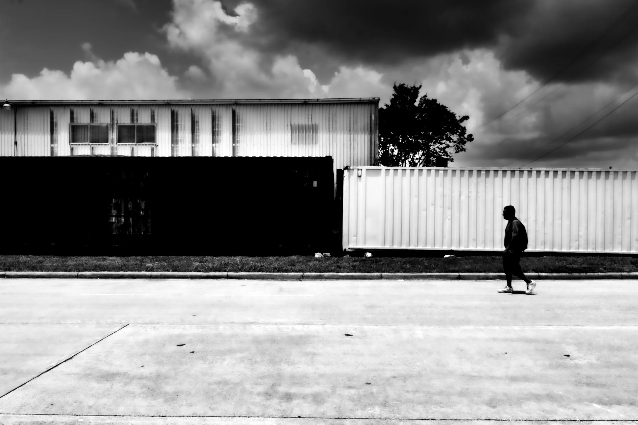 A person walking along a street with industrial buildings and a large container in the background, under a cloudy sky.