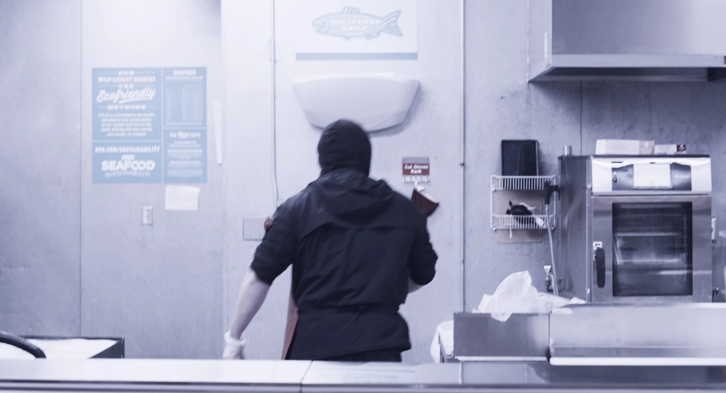 Person wearing black hoodie and apron working in a stainless steel kitchen, with appliances and posters on the wall.