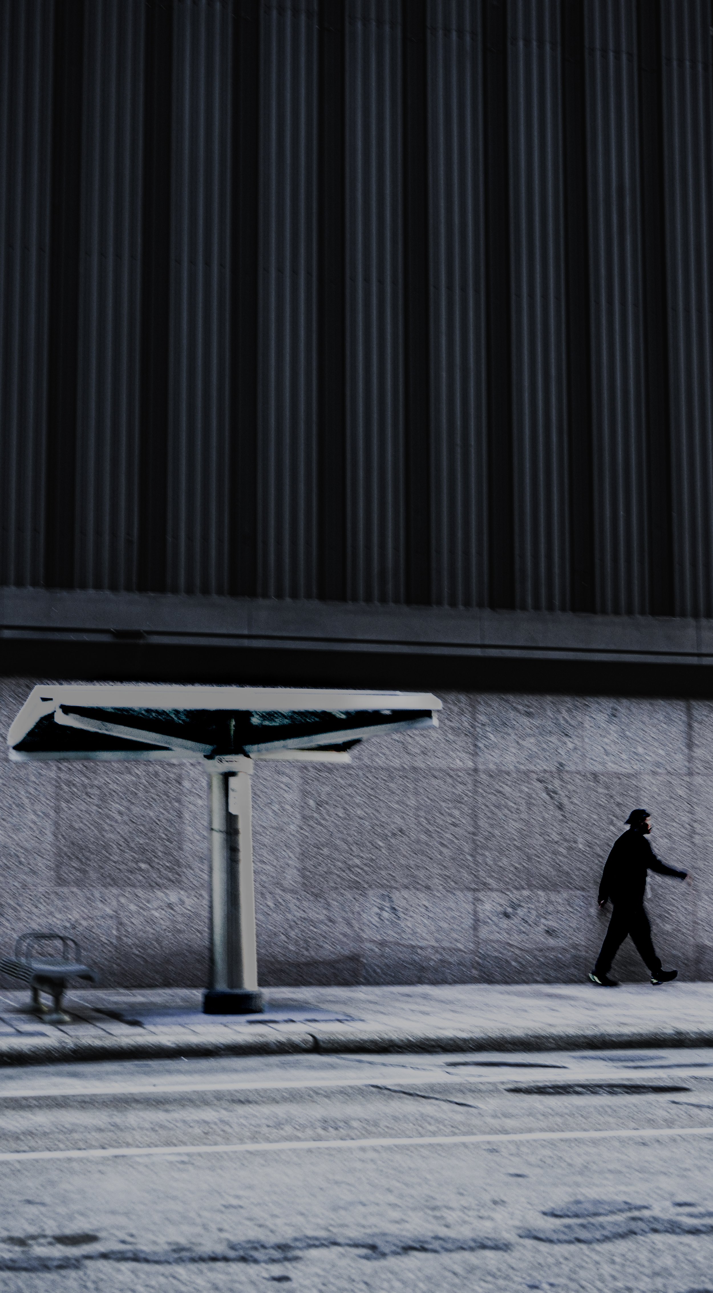 A person walking on a city sidewalk near a large modern building with a matte gray stone wall and vertical black siding. There is a large, tilted outdoor sign or artwork, a bench, and a street visible in the foreground.