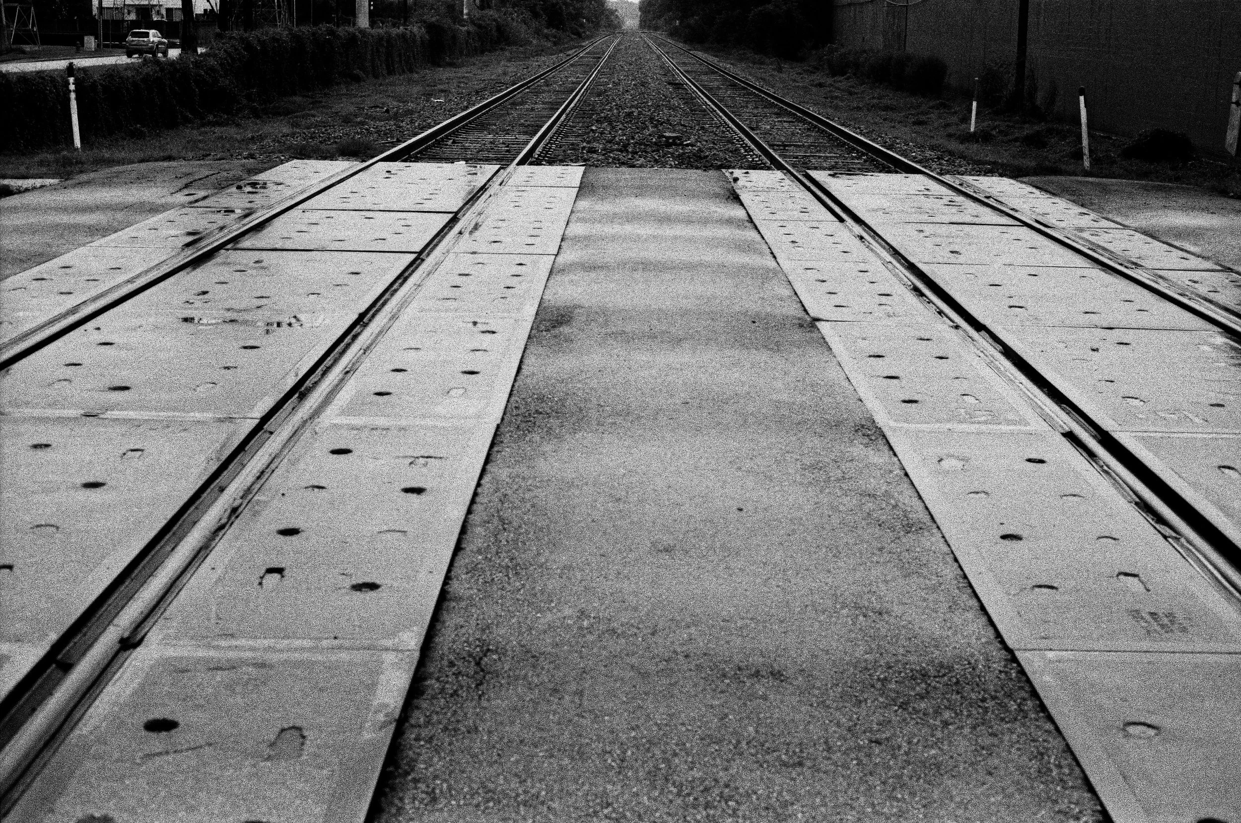 A black and white photo of a railway crossing with train tracks, crossing gate panels, and a paved road in the middle, extending into the distance with trees and cars on the side.
