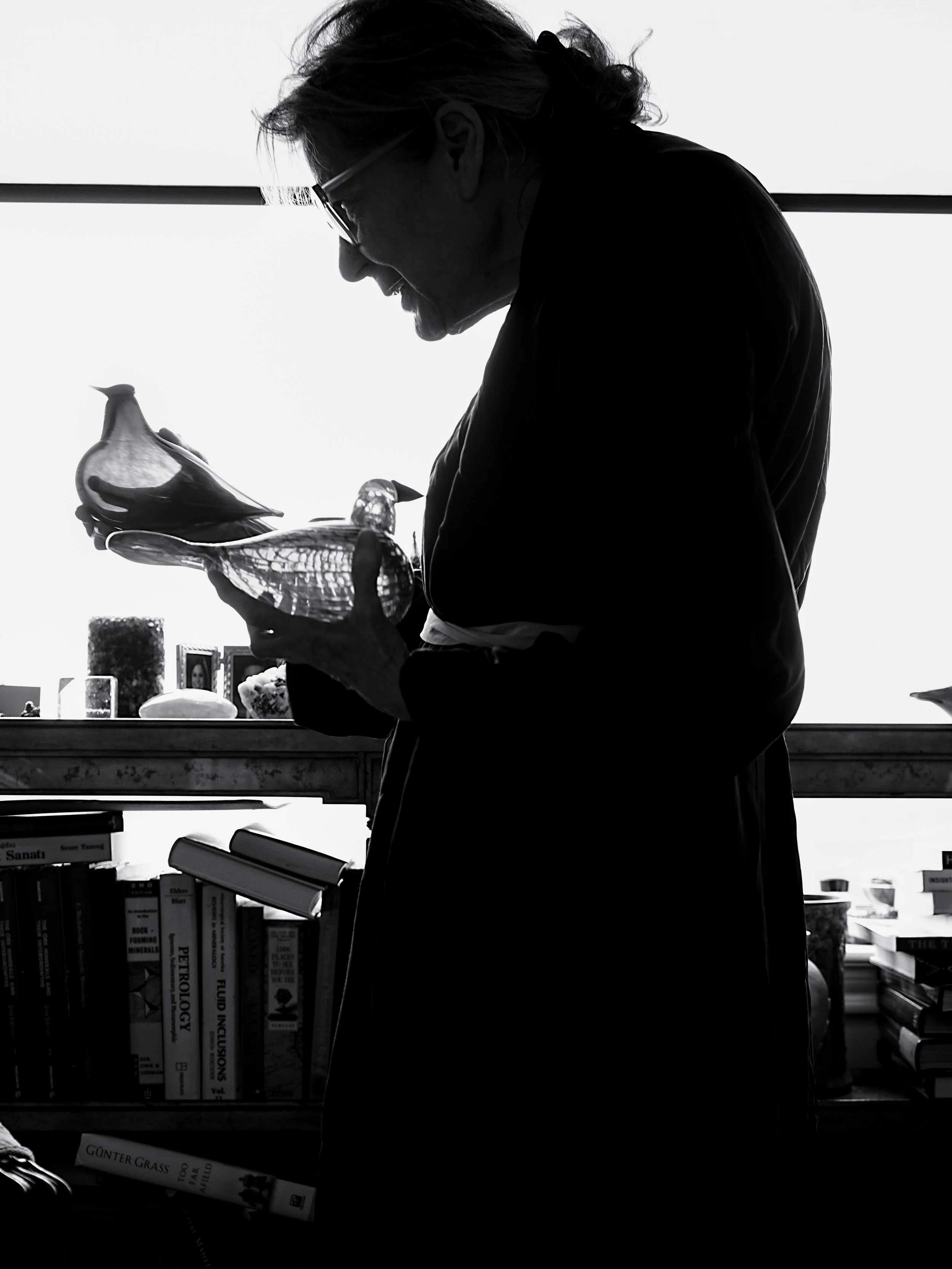 A woman in profile holding glass bird sculptures in front of a light background, with a bookshelf against the wall behind her.