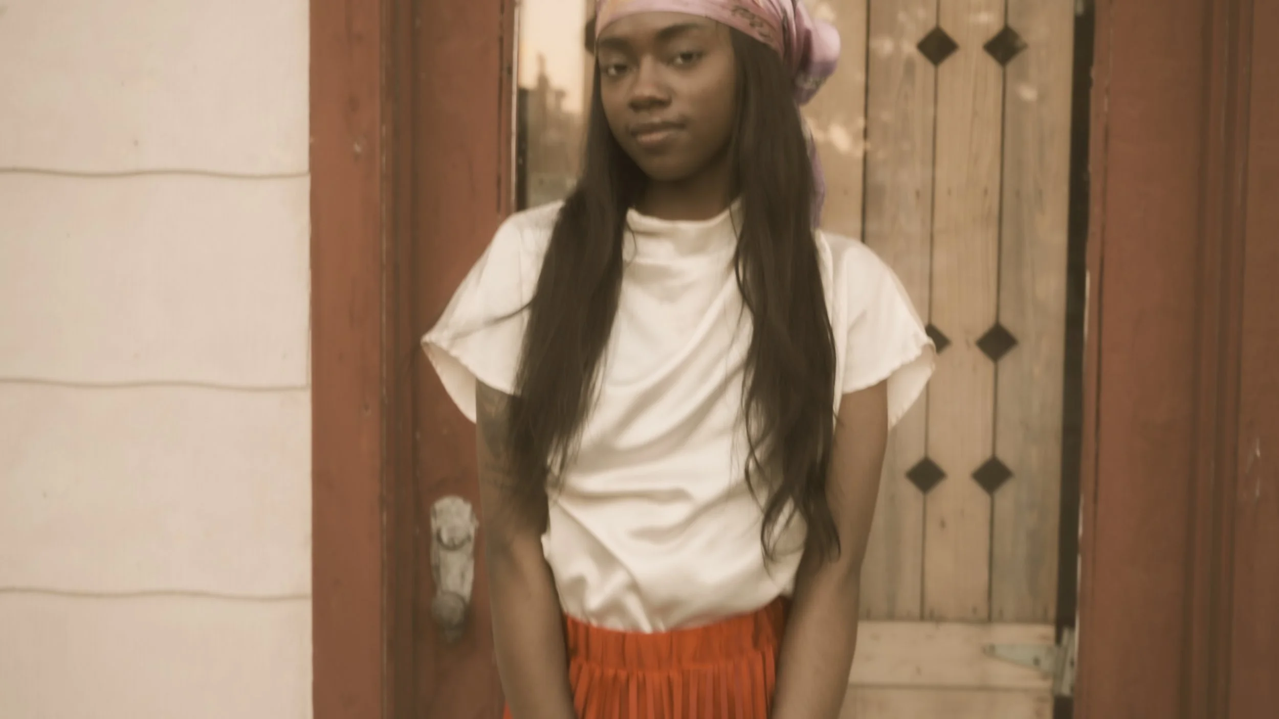 A young woman with long dark hair, wearing a light pink headscarf, white t-shirt, and orange skirt, standing in front of a wooden door.
