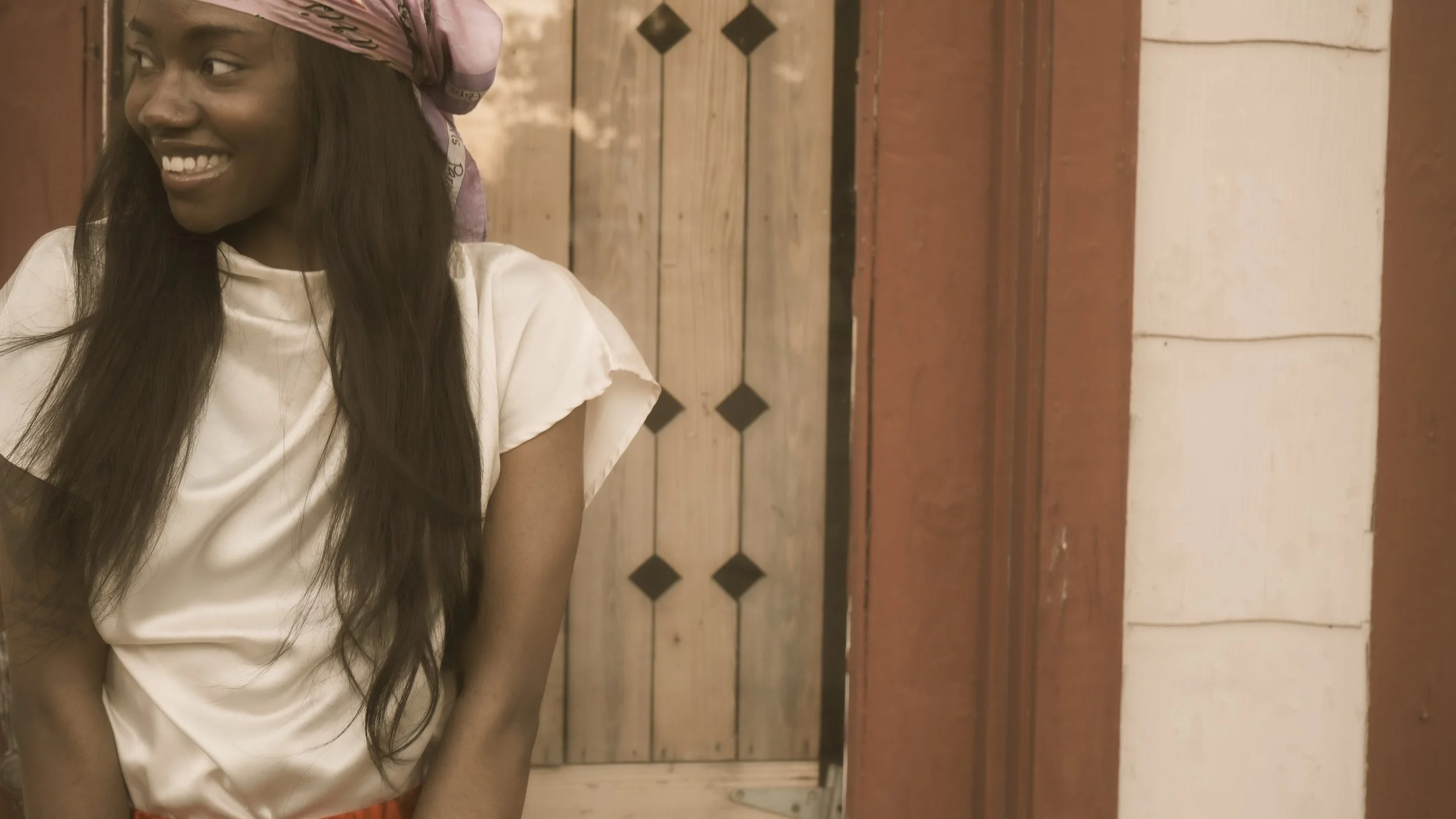 A woman with long dark hair wearing a pink headscarf and a white satin top, smiling and looking to her left, standing indoors near a wooden door with a rustic background.