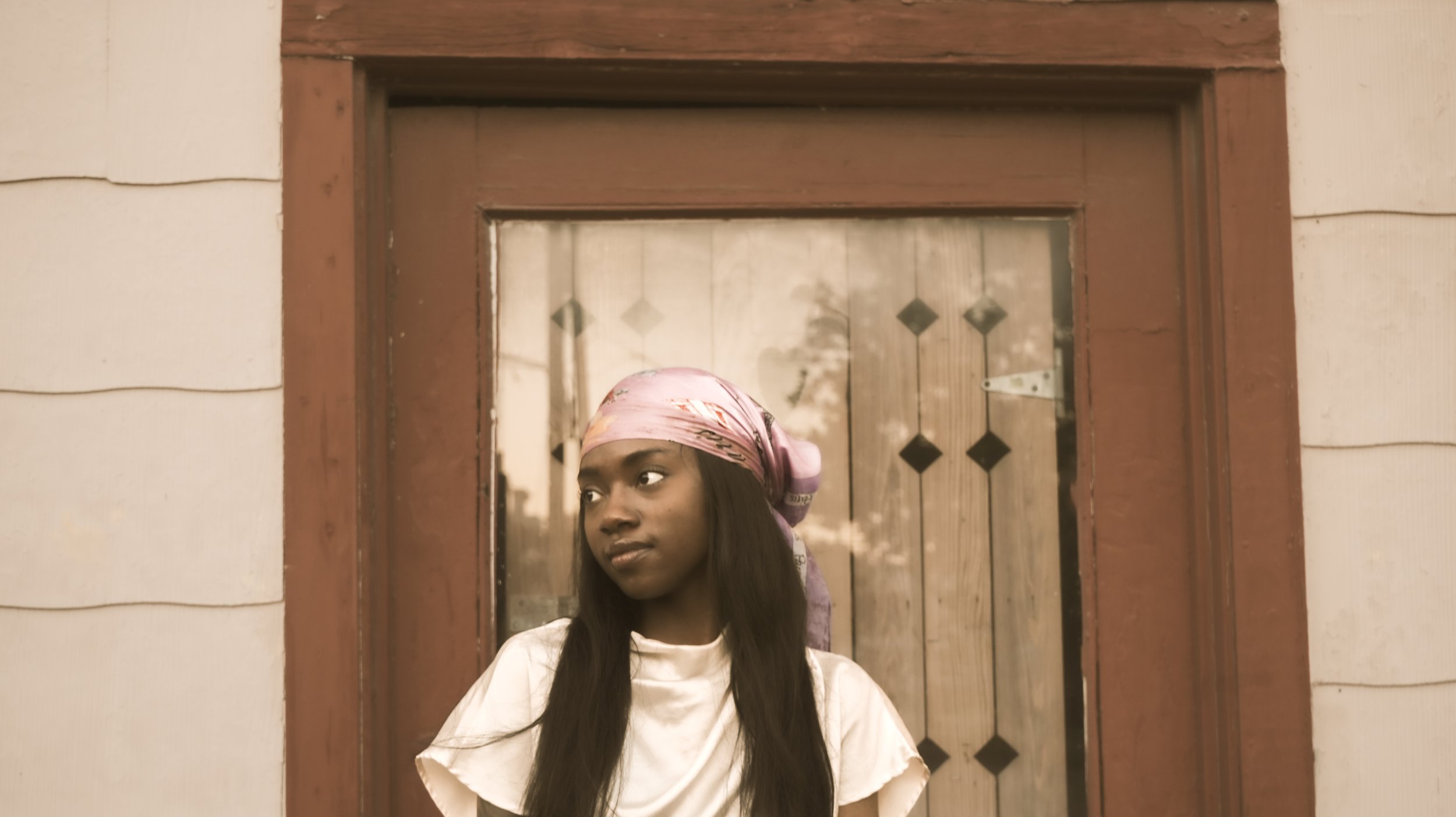 A young woman with long dark hair wearing a pink headscarf and cream-colored shirt, standing in front of a wooden door with glass panels.