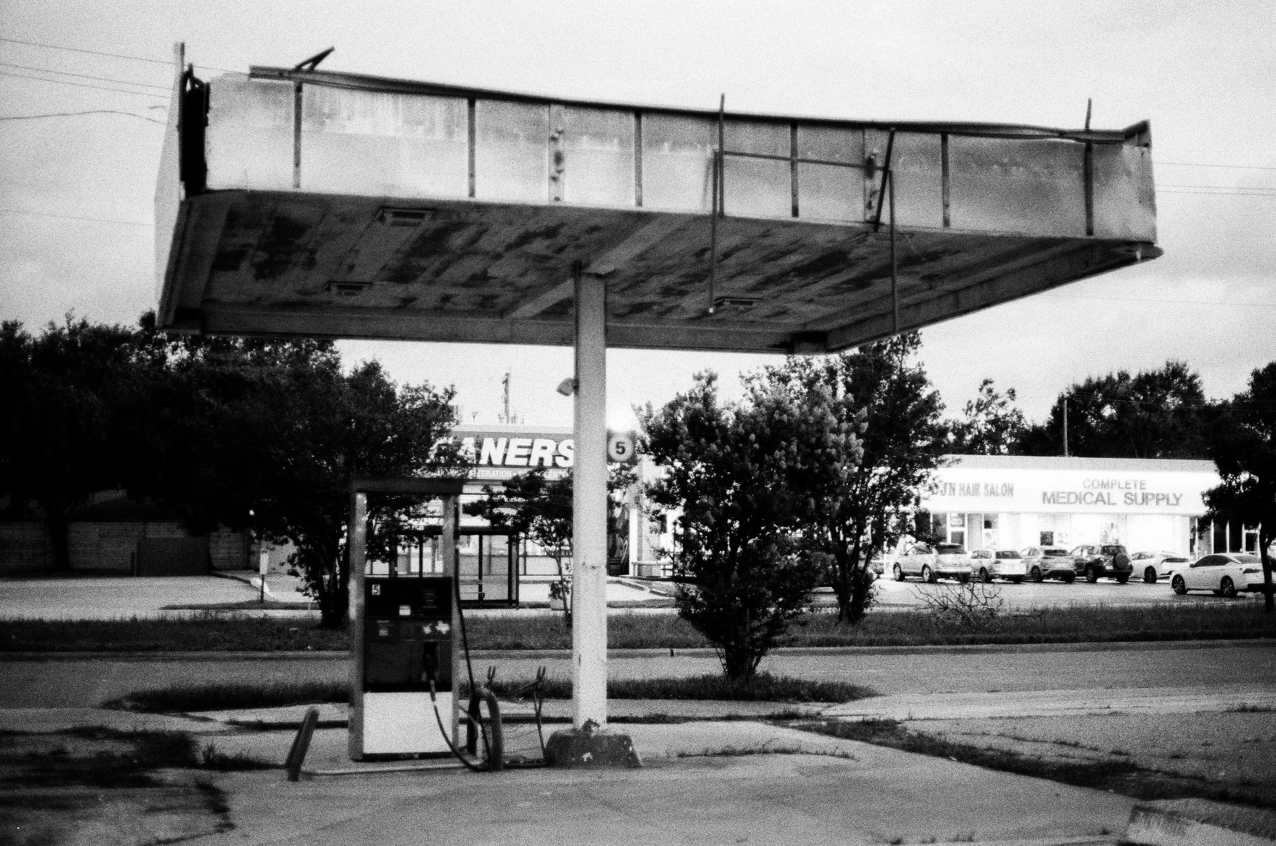 Black and white photo of an old, empty gas station with a single pump under a large, weathered canopy.