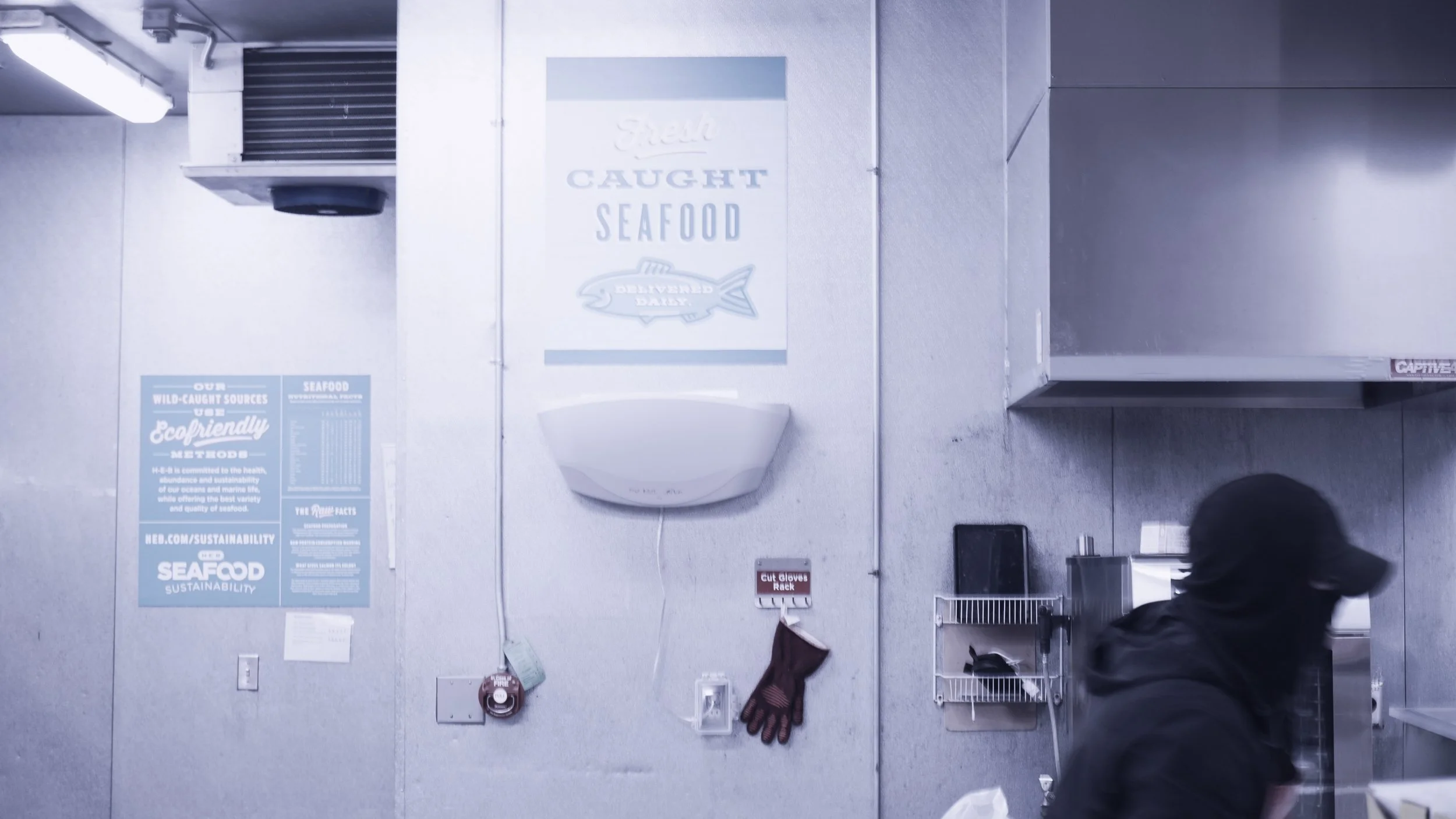 A person in black clothing and a cap walking in a commercial kitchen. The wall has a sign that reads "Caught Seafood" and an informational poster about seafood sustainability. There is a pair of gloves hanging on a hook, and various kitchen equipment