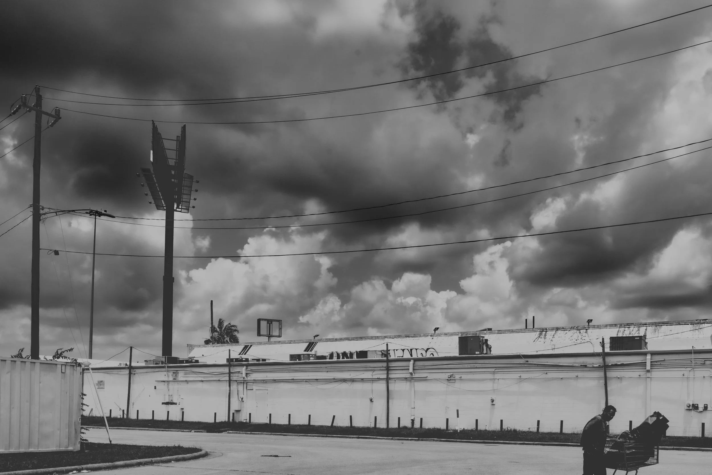 A person pushing a shopping cart near a white wall under a cloudy sky with utility poles and electrical wires.
