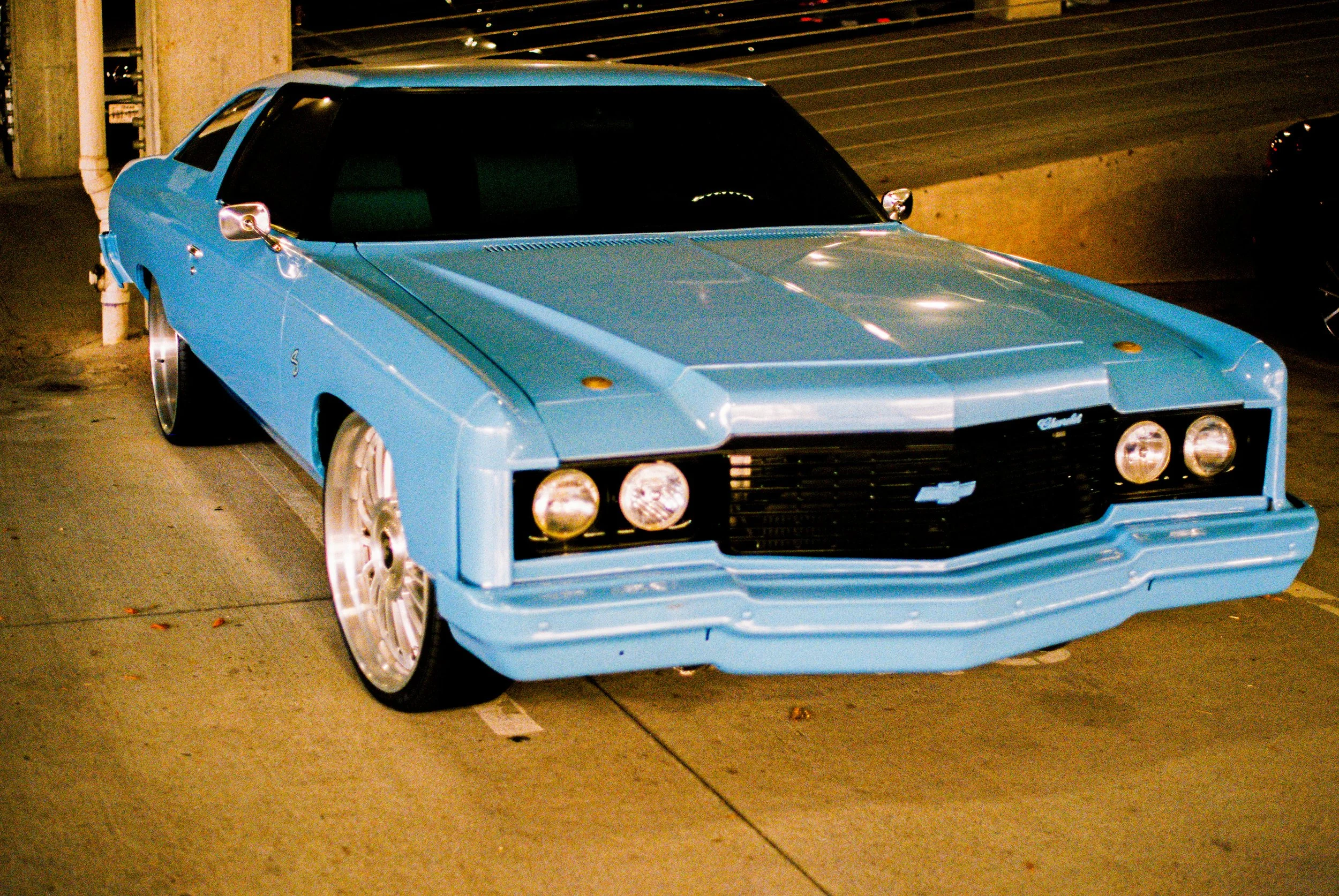 A vintage Chevrolet sports car painted in light blue, parked in a dimly lit indoor parking garage, featuring a sleek, angular design with a front grille and round headlights.