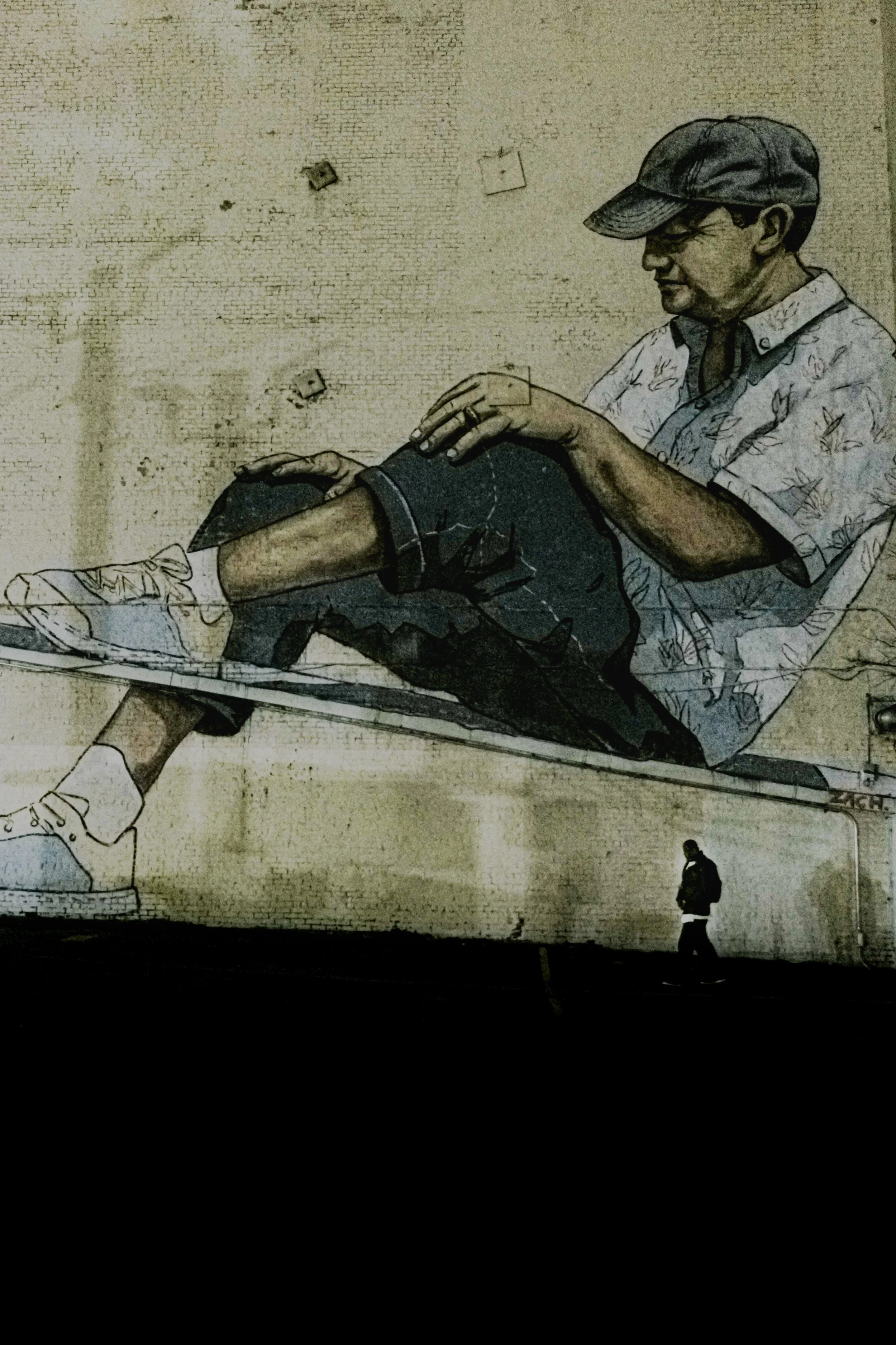 Street art mural of a man sitting, wearing a cap, with another person walking below on the sidewalk.