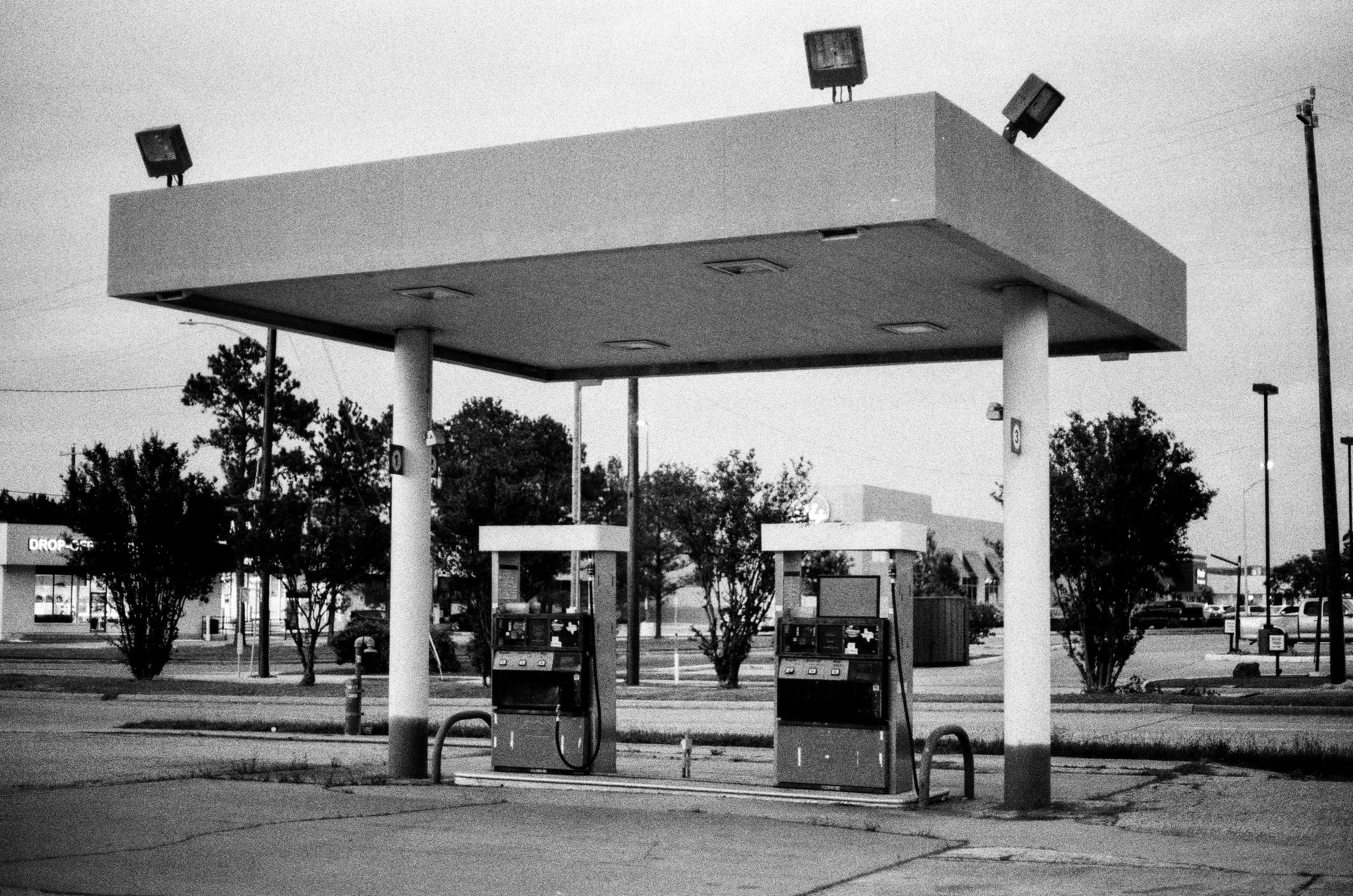 A black and white photo of an empty gas station with two fuel pumps under a large flat canopy, with trees and a parking lot in the background.