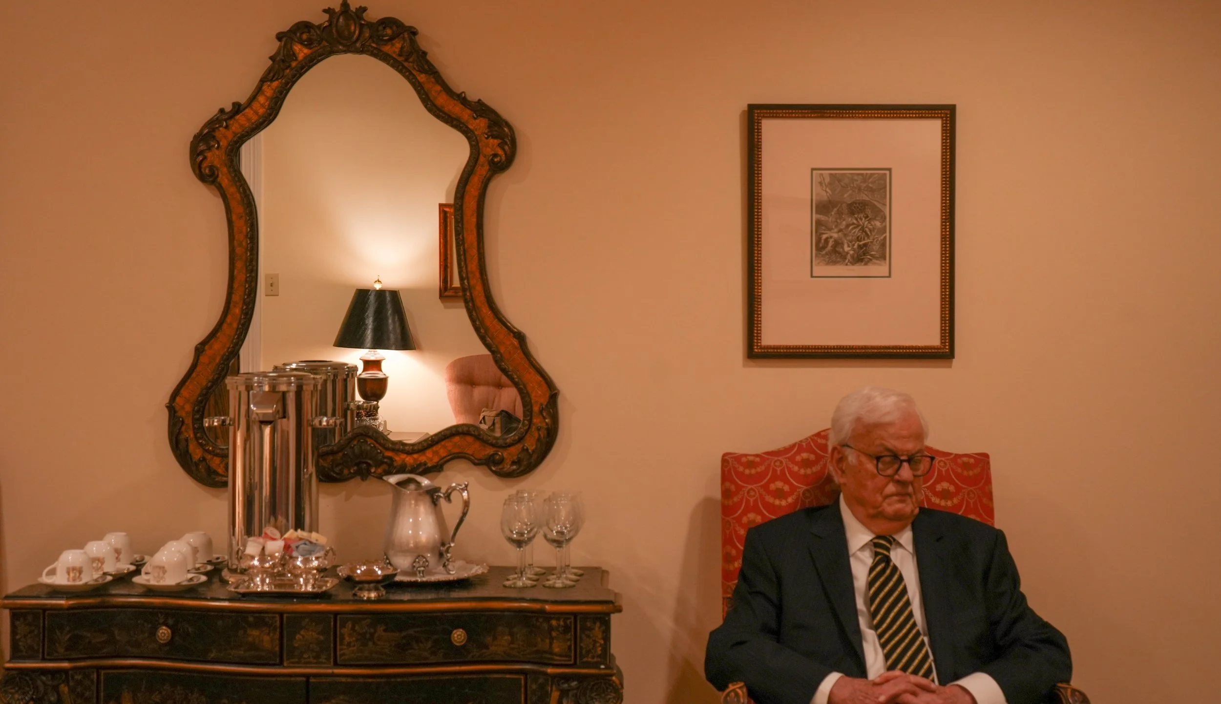 An elderly man wearing glasses and a suit sits with his hands clasped on a table in a living room with a decorative mirror, a lamp, a framed picture, and a wooden sideboard with cups, a tea set, and glasses.
