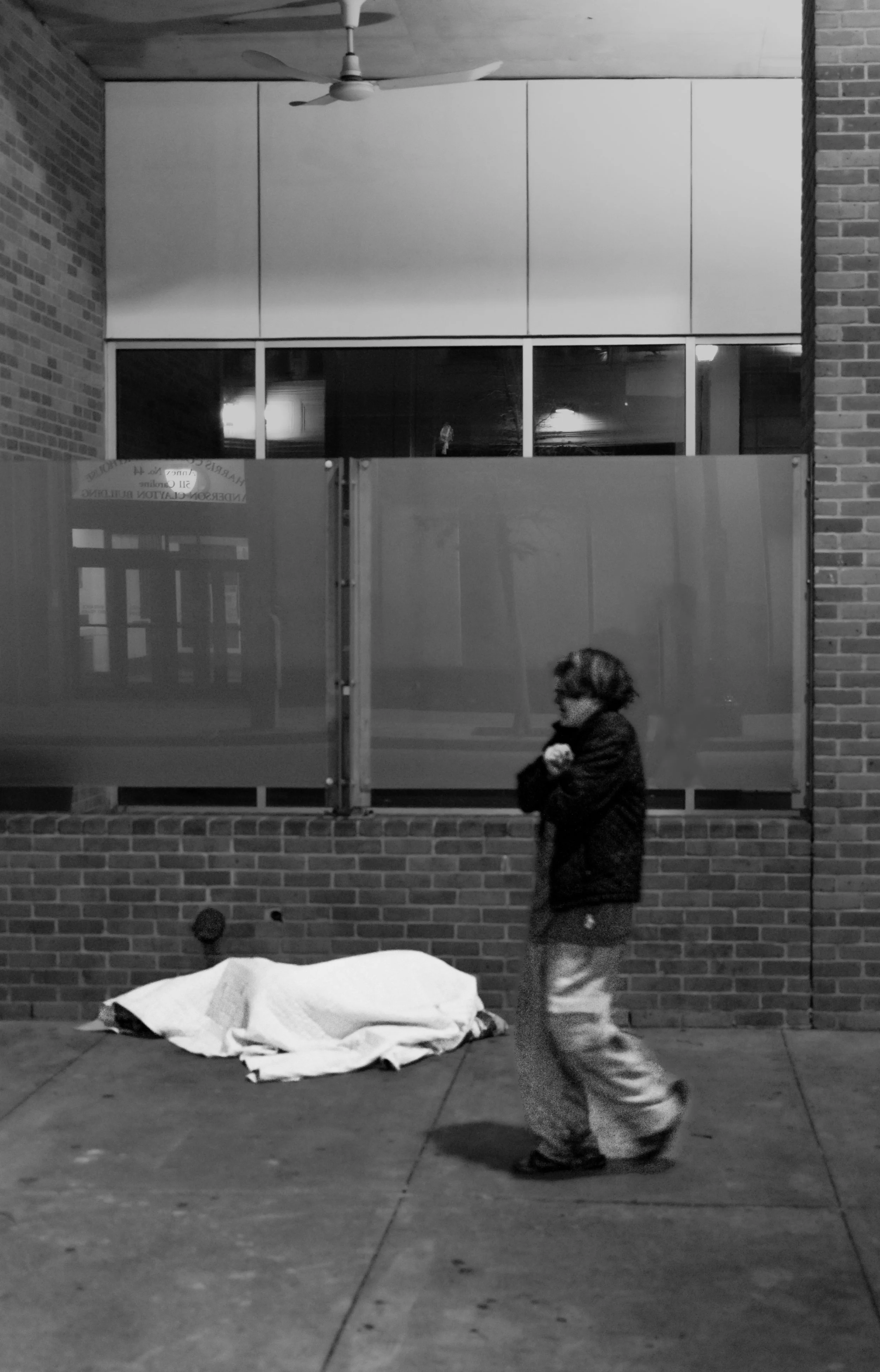 A person walking past a covered body lying on the sidewalk outside a building. The scene is in black and white, with the person wearing glasses, a jacket, and sweatpants, and appears to be at night.
