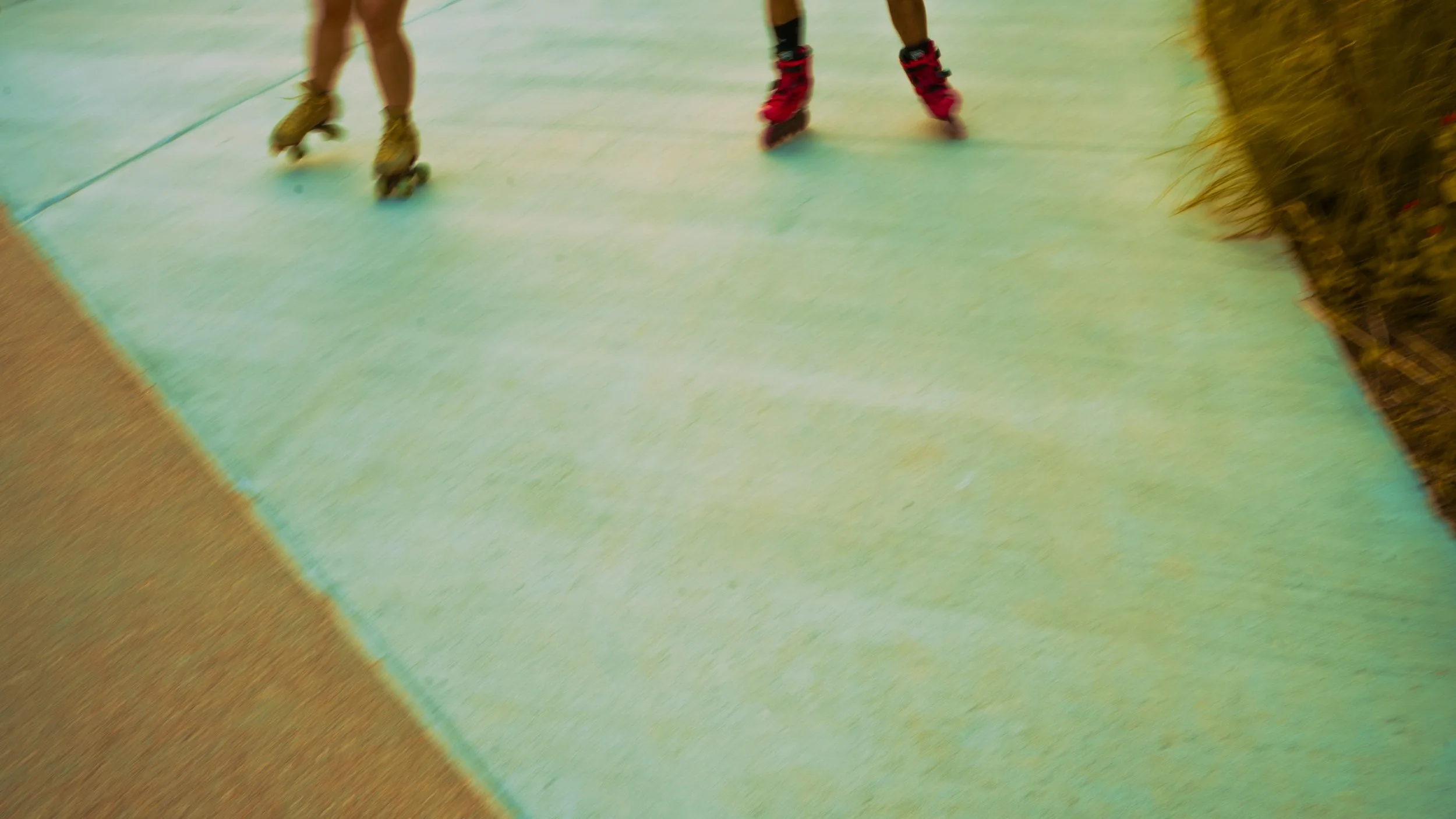 Two people rollerblading on a green indoor surface, with one wearing tan skates and the other in pink skates.