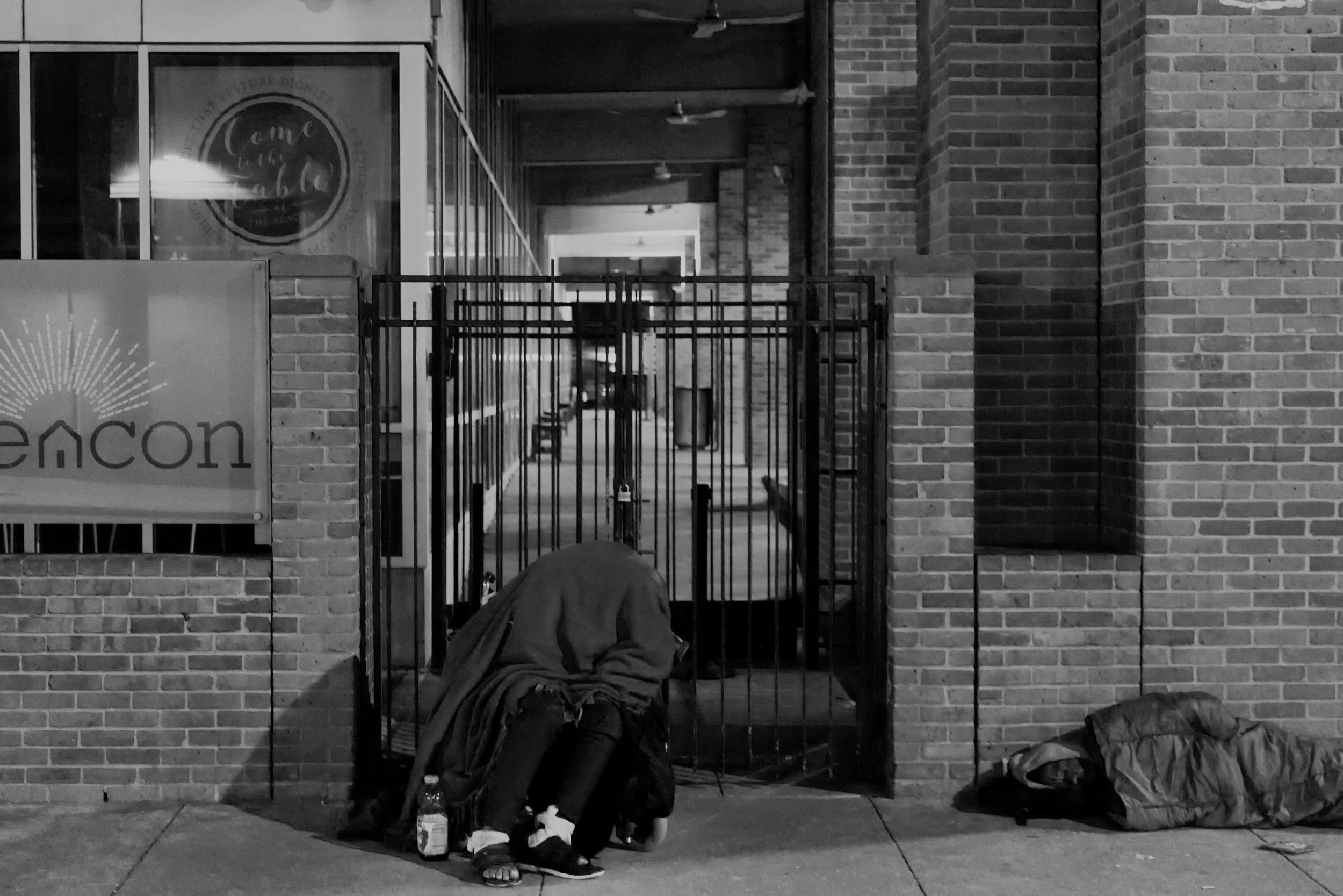 A person sitting on the sidewalk with a blanket over their head, next to a bottle and a bag, outside a building with a gated entrance and a sign that says "encon." The person appears to be sleeping or resting.