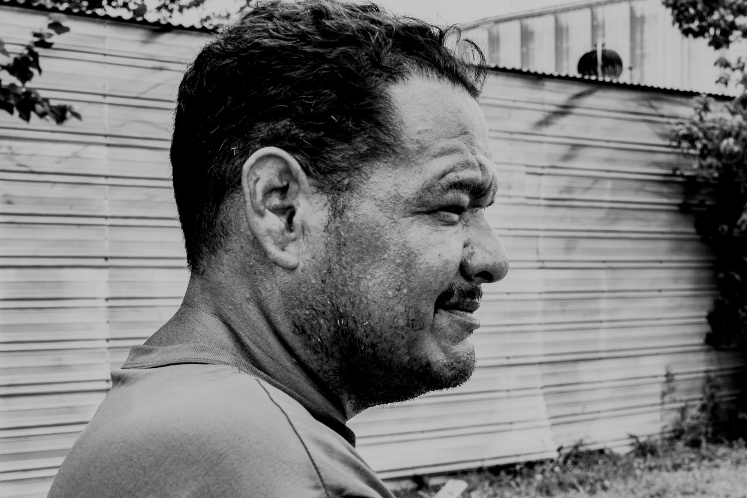 A close-up side profile of a man with short, dark hair, sitting outdoors in front of a wooden fence, with visible sweat and dirt on his face.