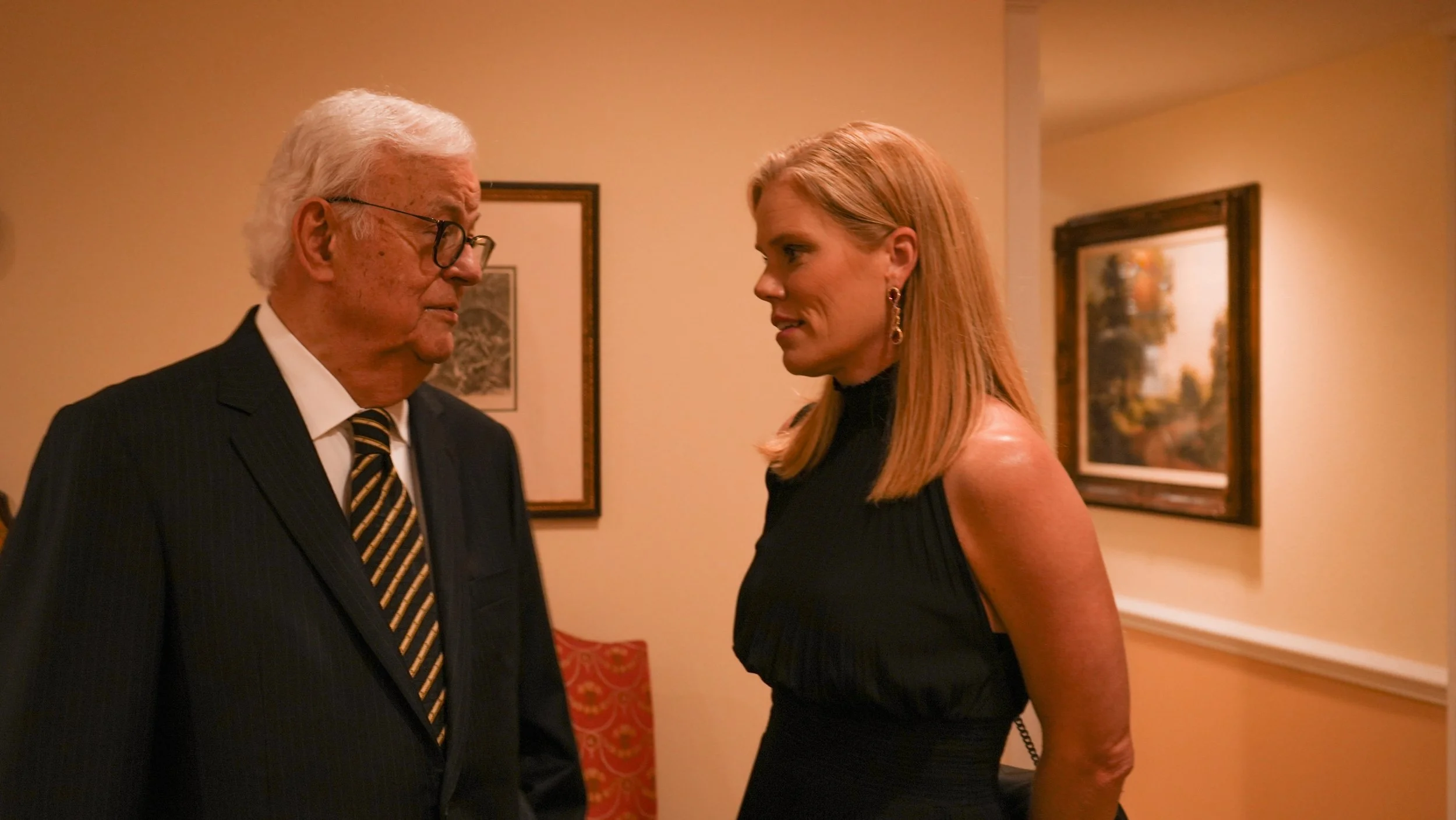 An older man in a suit and tie, wearing glasses, and a woman in a black sleeveless dress with earrings, facing each other and talking in a warmly lit room with framed art on the walls.