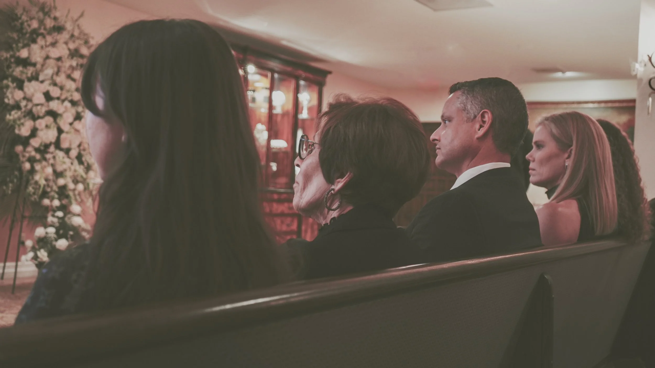 People sitting in pews at a funeral service or memorial, with floral arrangements in the background.