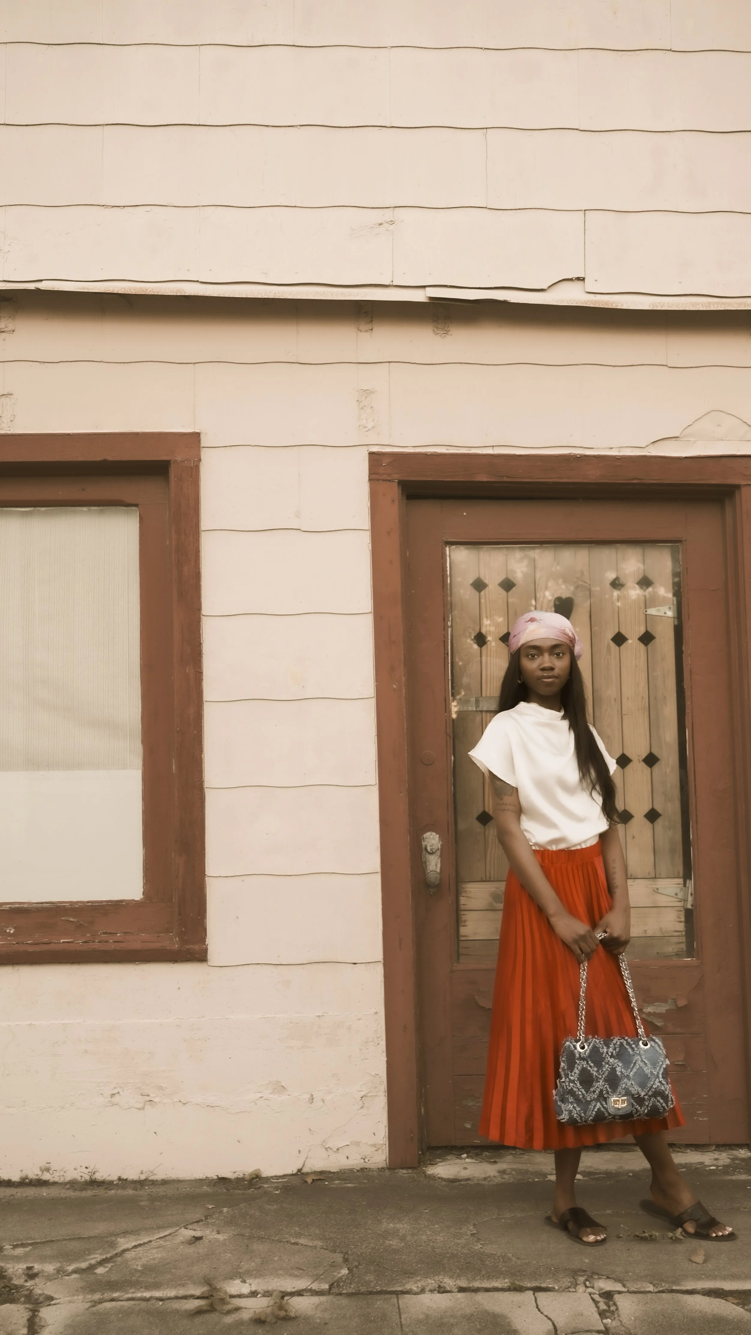 A woman standing outside a wooden door with a window, wearing a white top, a red striped skirt, a pink headscarf, brown sandals, and carrying a snakeskin patterned handbag.