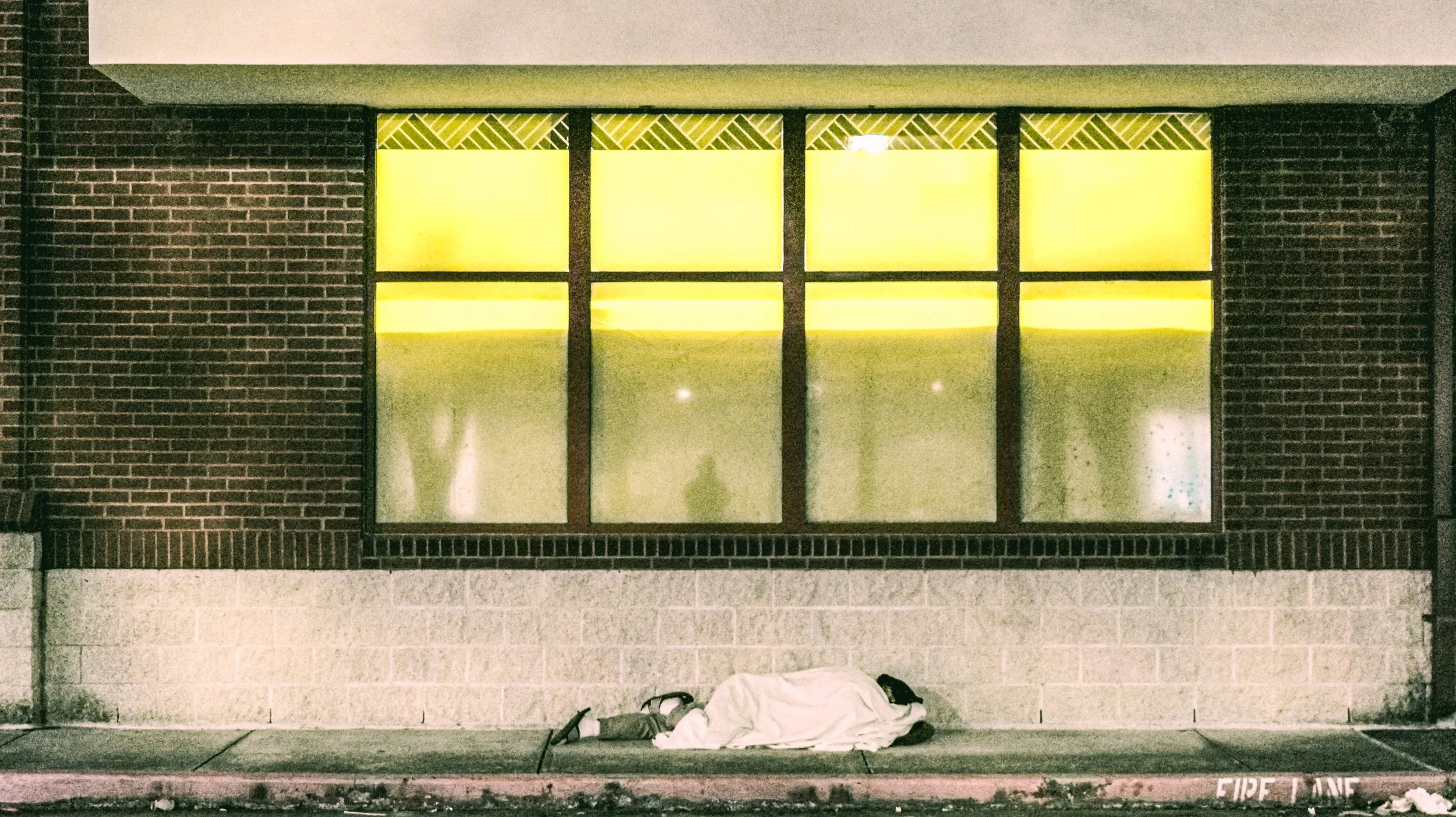 A person lying on the sidewalk in front of a building with large yellow-lit windows, covered with a white sheet, with a handbag and other items next to them behind the sheet, and a fire lane painted on the curb.