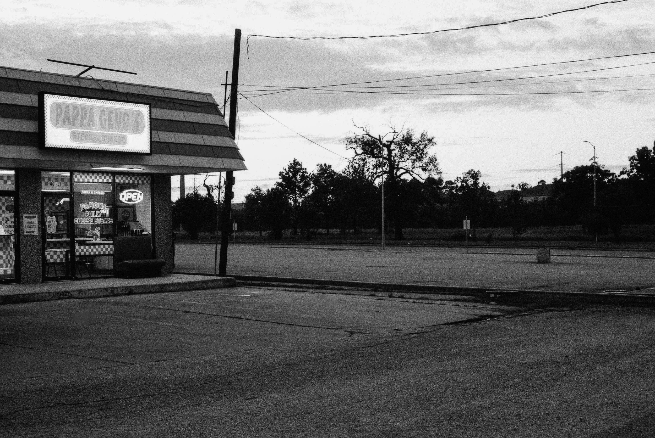 A black-and-white photo of a small fast food restaurant named Pappa Gino's, with a sign that says 'Steak & Cheese'. The restaurant has a checkered pattern on the lower part of the exterior walls and a neon 'Open' sign in the window. There are trees a