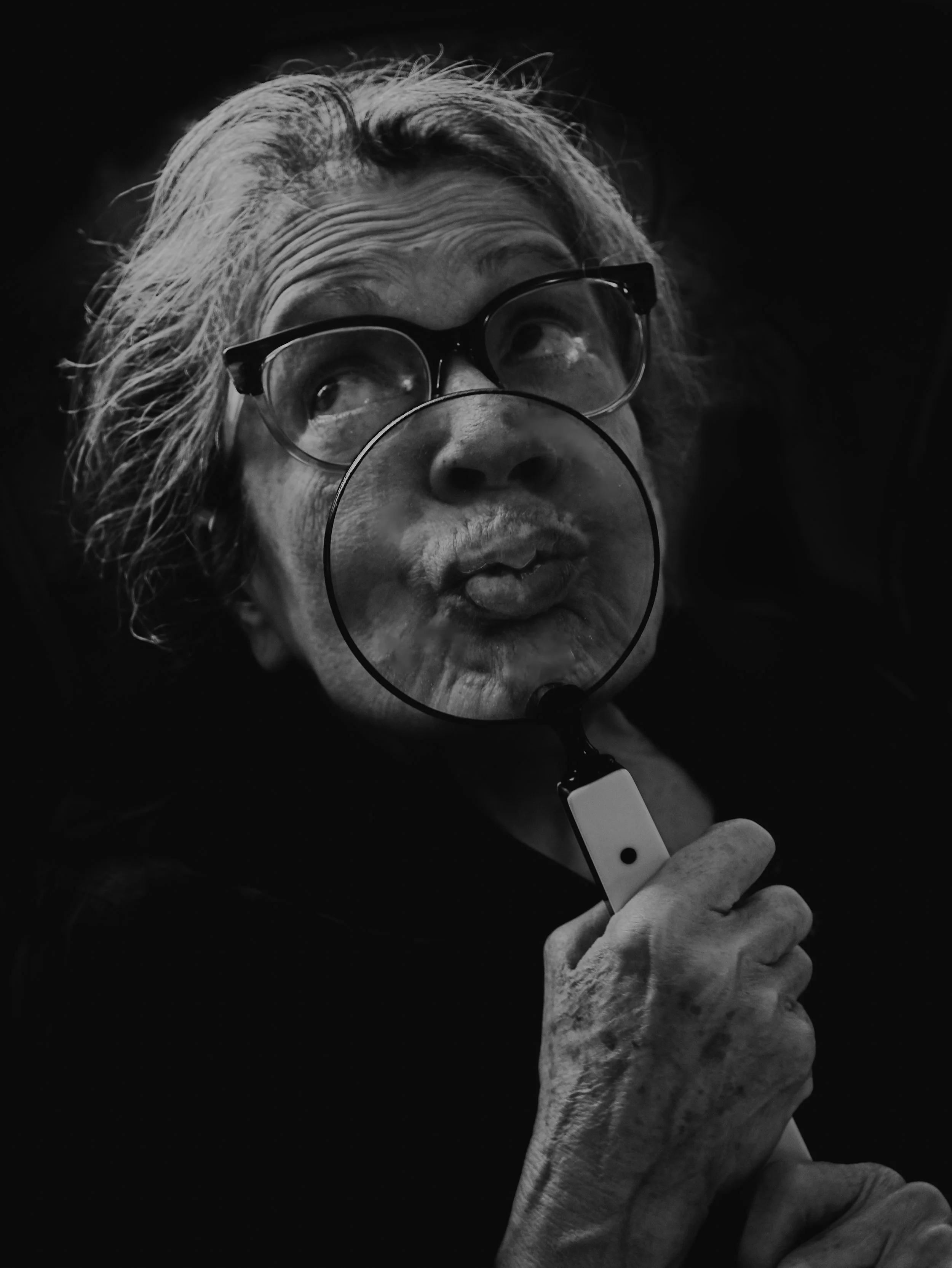 An elderly woman with gray hair and glasses holding a magnifying glass in front of her lips, which are puckered as if blowing a kiss, in black and white.