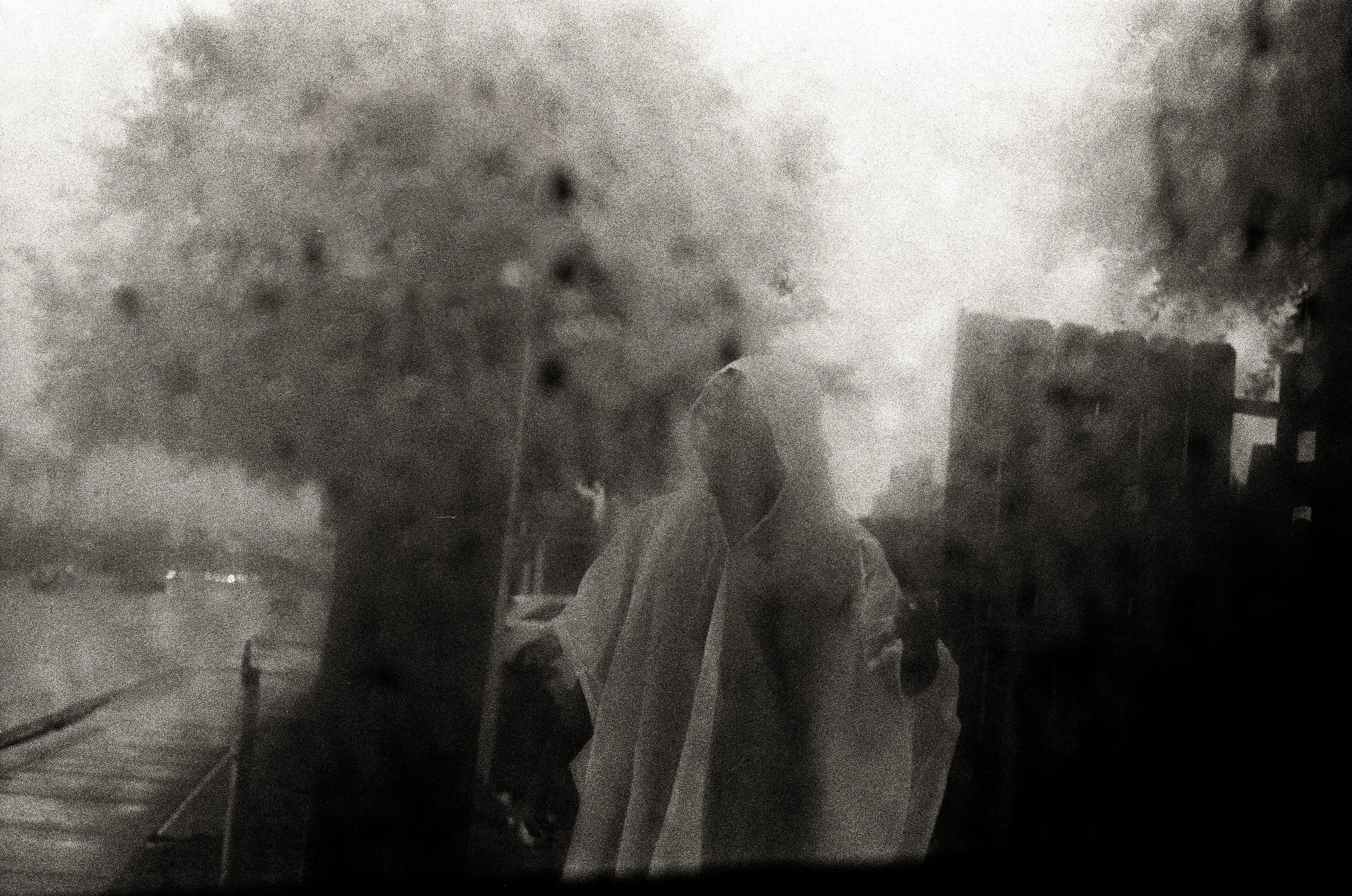 Black and white photograph of two people sharing a close, intimate moment on the street, with onlookers and a building in the background, viewed through a rain-covered window.