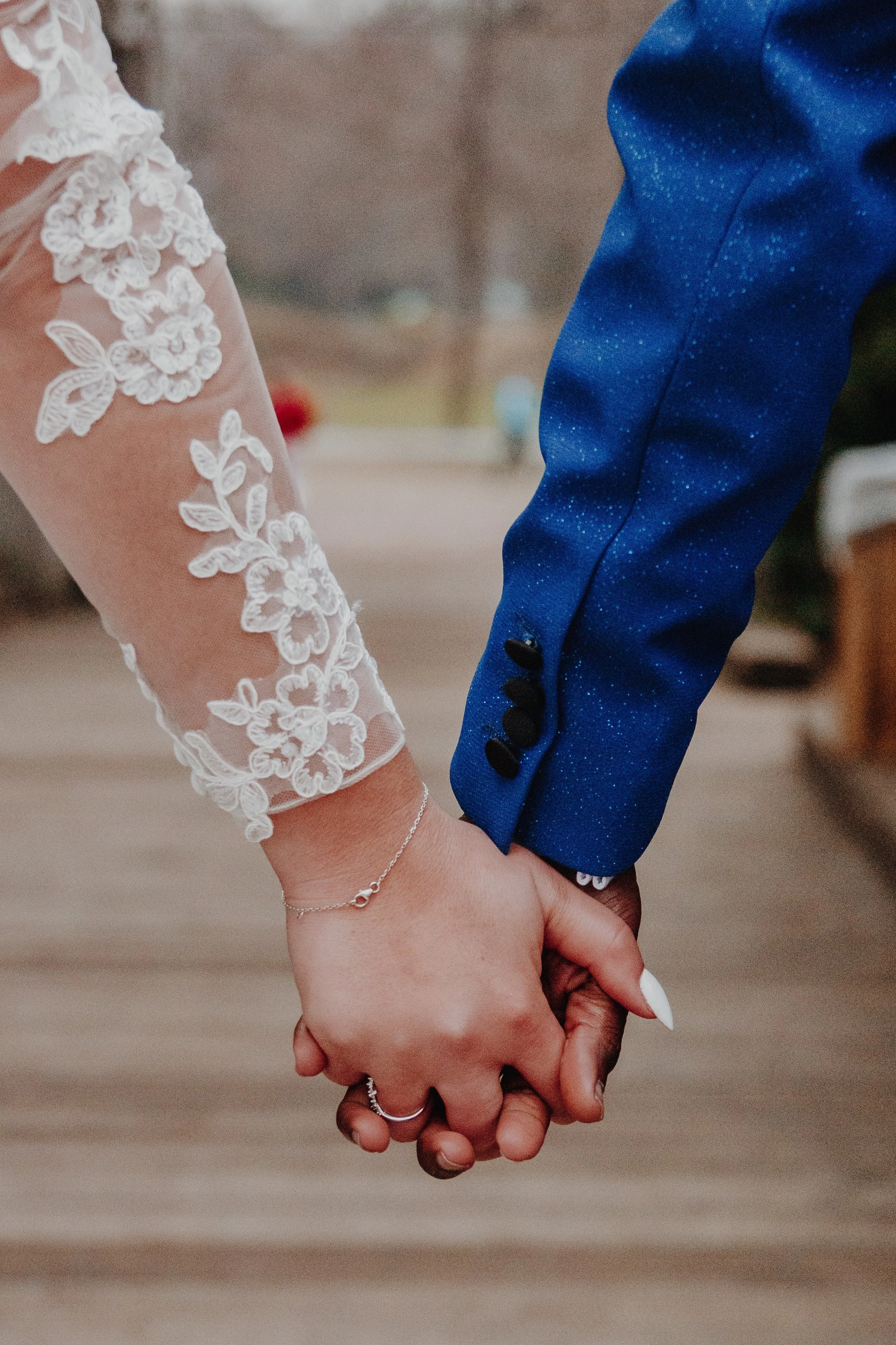 A bride and groom holding hands, with the bride wearing a white lace sleeve and the groom in a blue suit.