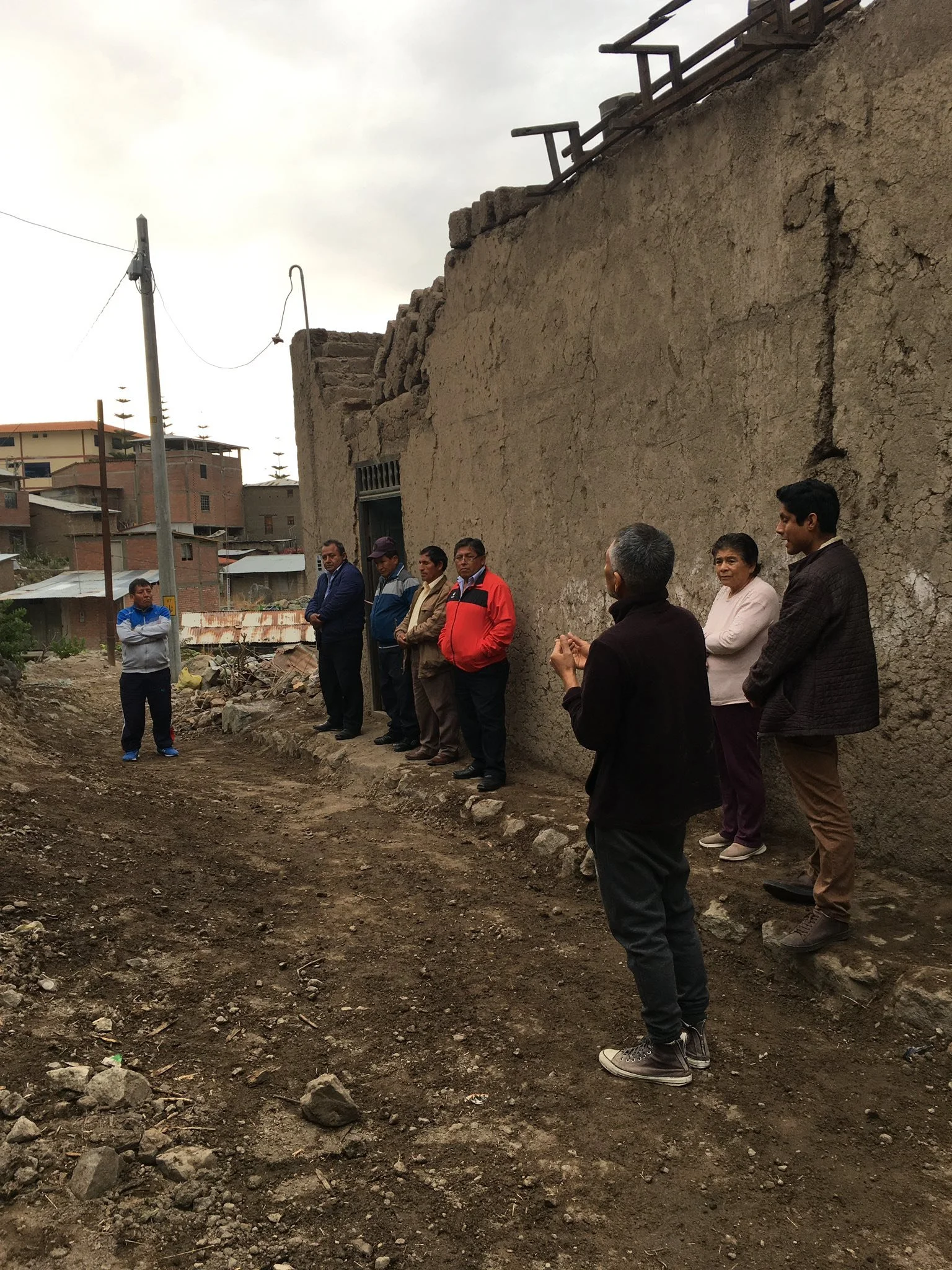 Grupo de personas dialogando al aire libre en una calle de tierra frente a un muro de adobe en un barrio popular.
