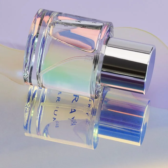 A perfume bottle with iridescent reflections rests on its side against a light purple background. The shiny metallic cap is prominent, and the bottle's surface displays a spectrum of soft colors.