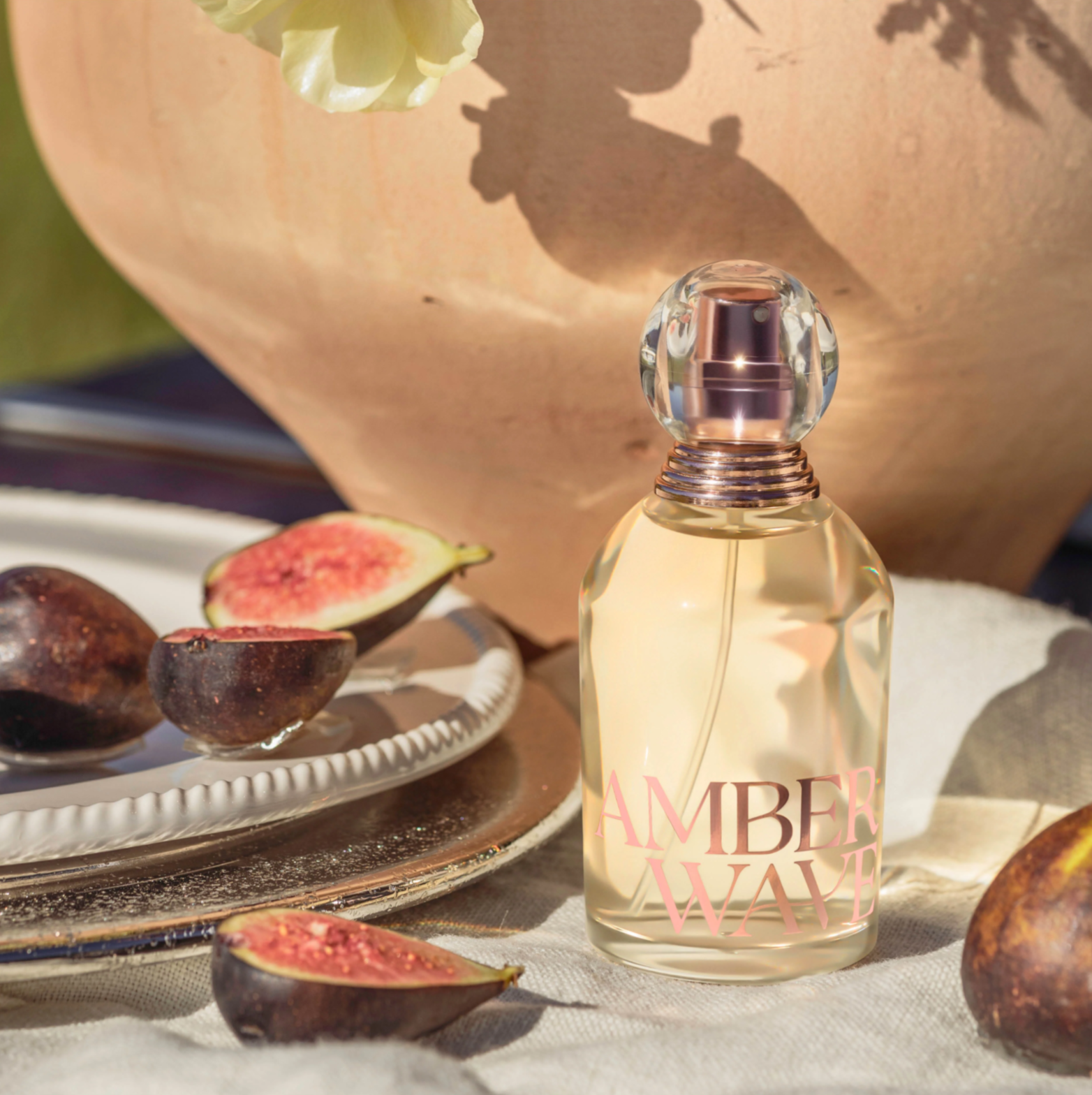 A perfume bottle with Amber Waves sits on a beige cloth. Cut figs on a white plate and a large terracotta vase with shadows are in the background. Warm sunlight enhances the scene, giving it an elegant, natural feel.