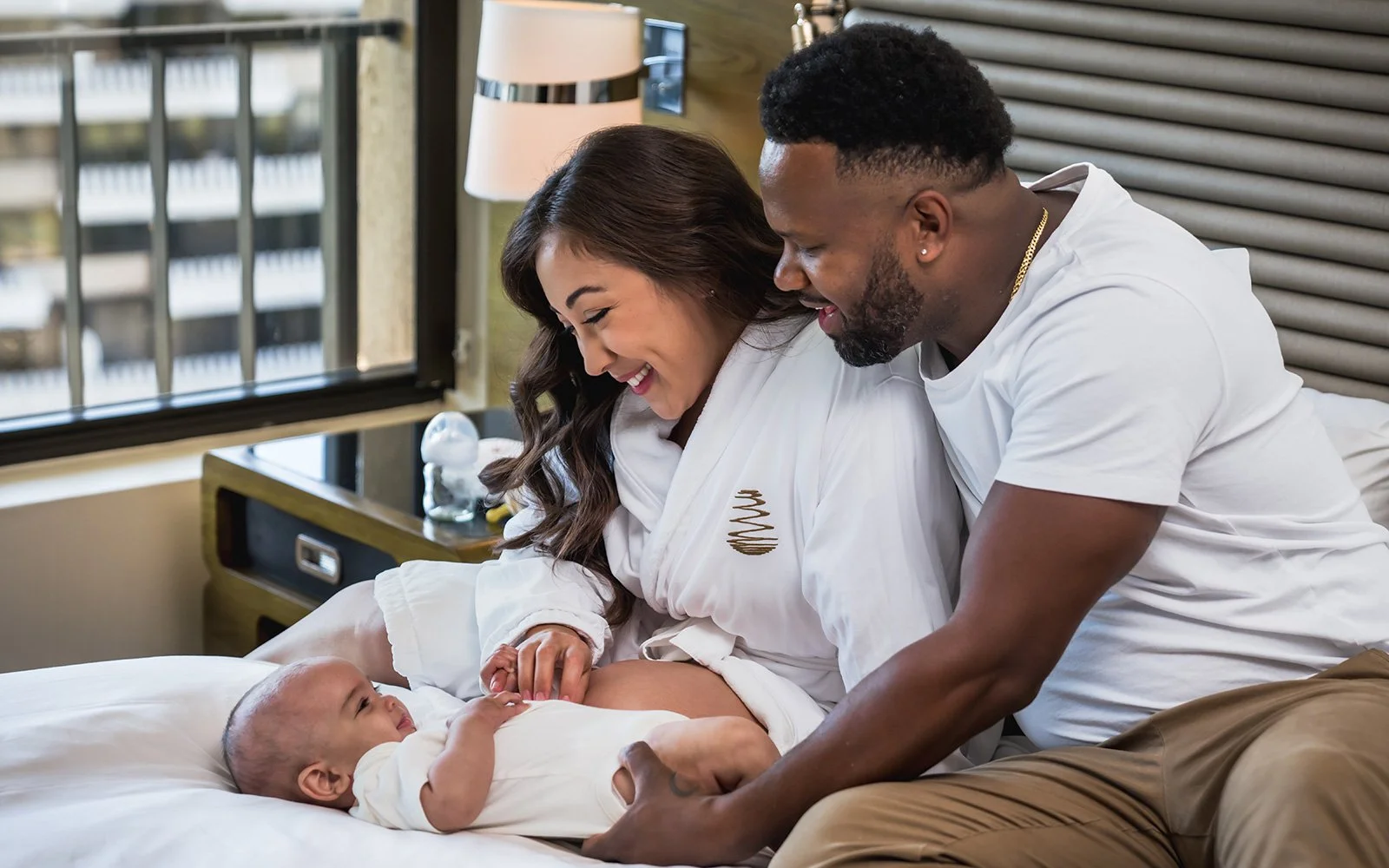 A joyful couple in a warmly lit bedroom smiles at their baby lying on a bed. The mother wears a white robe, and the father is in a white shirt.