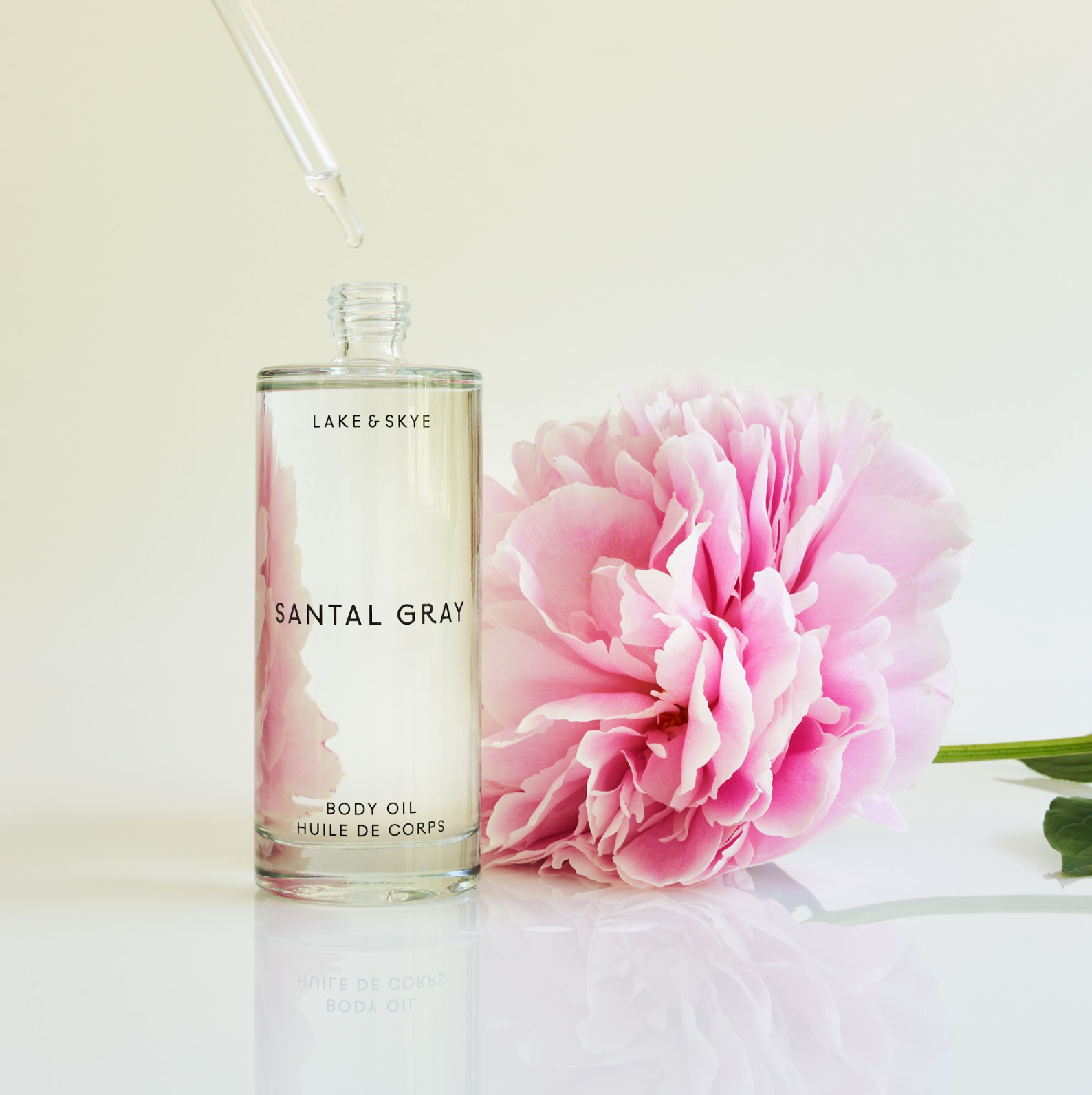 A clear glass bottle labeled Lake & Skye Santal Gray Body Oil stands next to a large pink peony bloom. A dropper is inserted into the bottle. The background is a soft, neutral white.