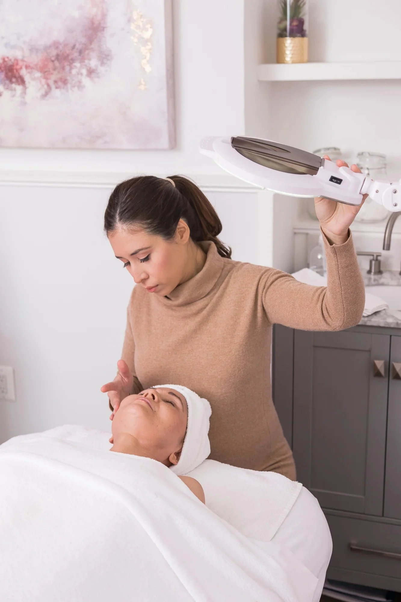 A woman in a beige turtleneck provides a facial treatment to a person lying on a spa bed. The ambiance is calm with soft, warm lighting.