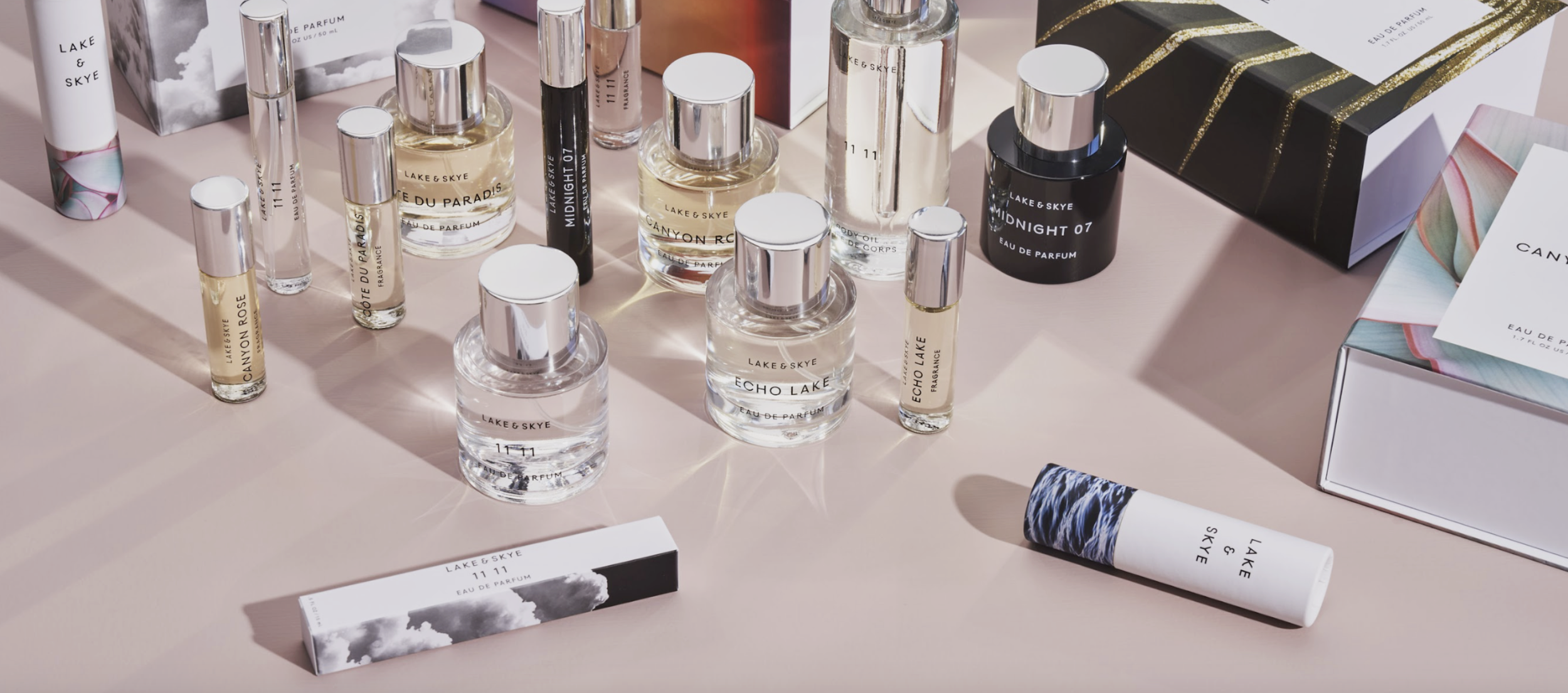 A variety of Lake & Skye perfume bottles and boxes are displayed on a light pink surface. The bottles, in different sizes, contain transparent liquids. The packaging features minimalist designs with sleek fonts and subtle color accents.