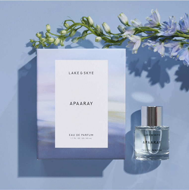 Perfume bottle and box labeled Lake & Skye Apaaray on a blue surface, with delicate flowers in the background casting shadows.