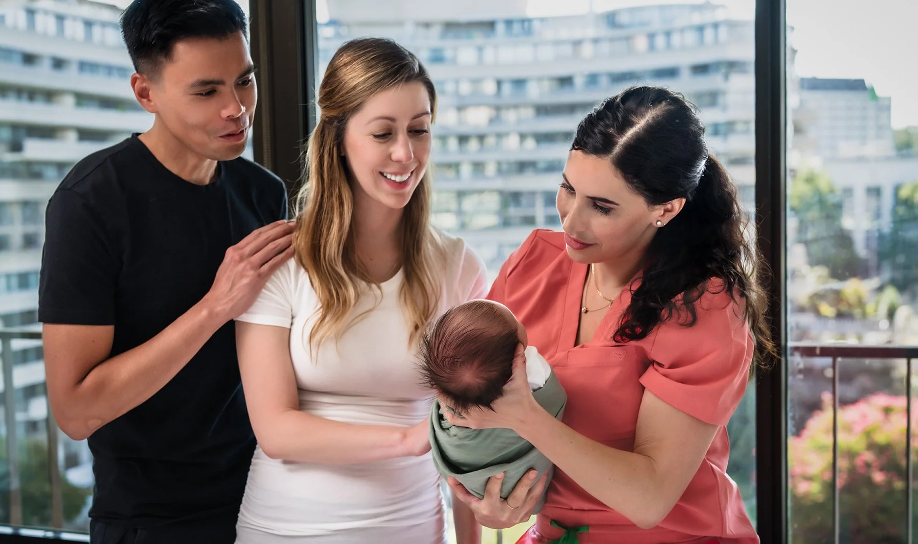 A couple smiles as a nurse in a pink uniform gently holds their newborn wrapped in a green blanket. Bright window view of urban buildings outside.