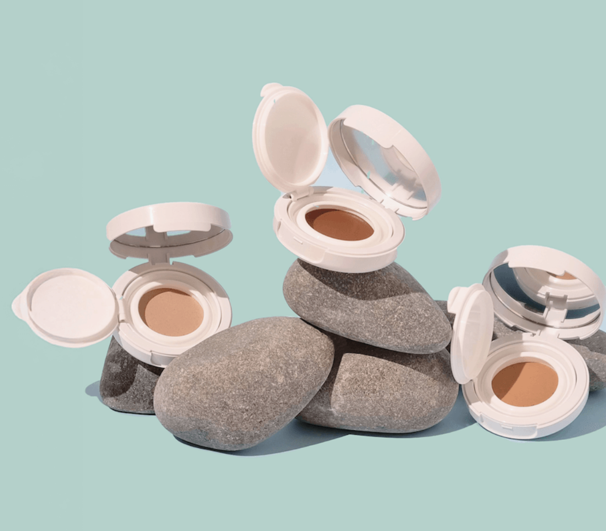 Open white compact cases with makeup sit on stacked gray stones against a light aqua background, conveying a serene and natural aesthetic.