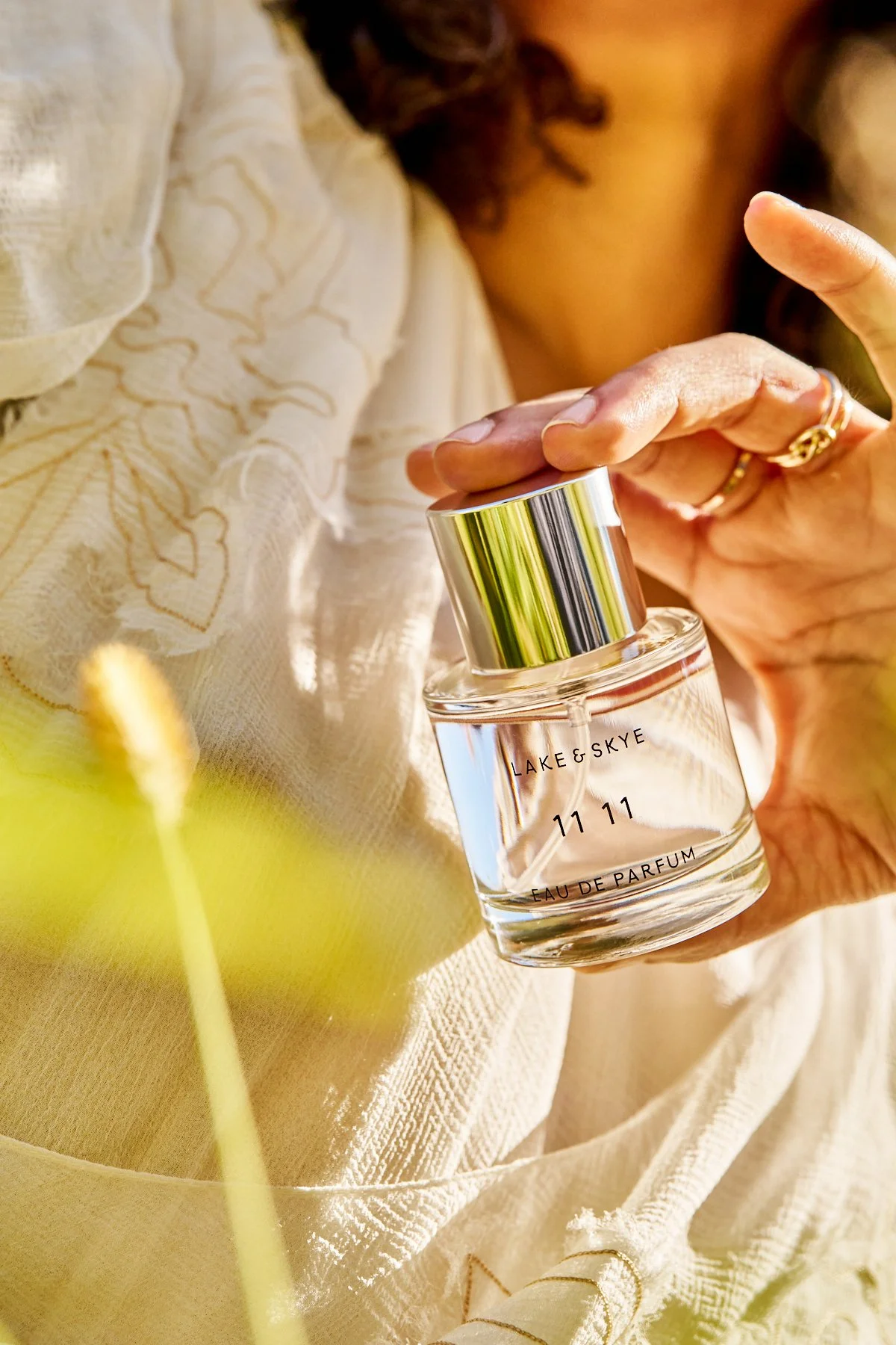 Close-up of a person holding a perfume bottle labeled Lake & Skye 11 11 Eau de Parfum. The person wears a white textured outfit, and the background features green leaves, suggesting an outdoor setting.