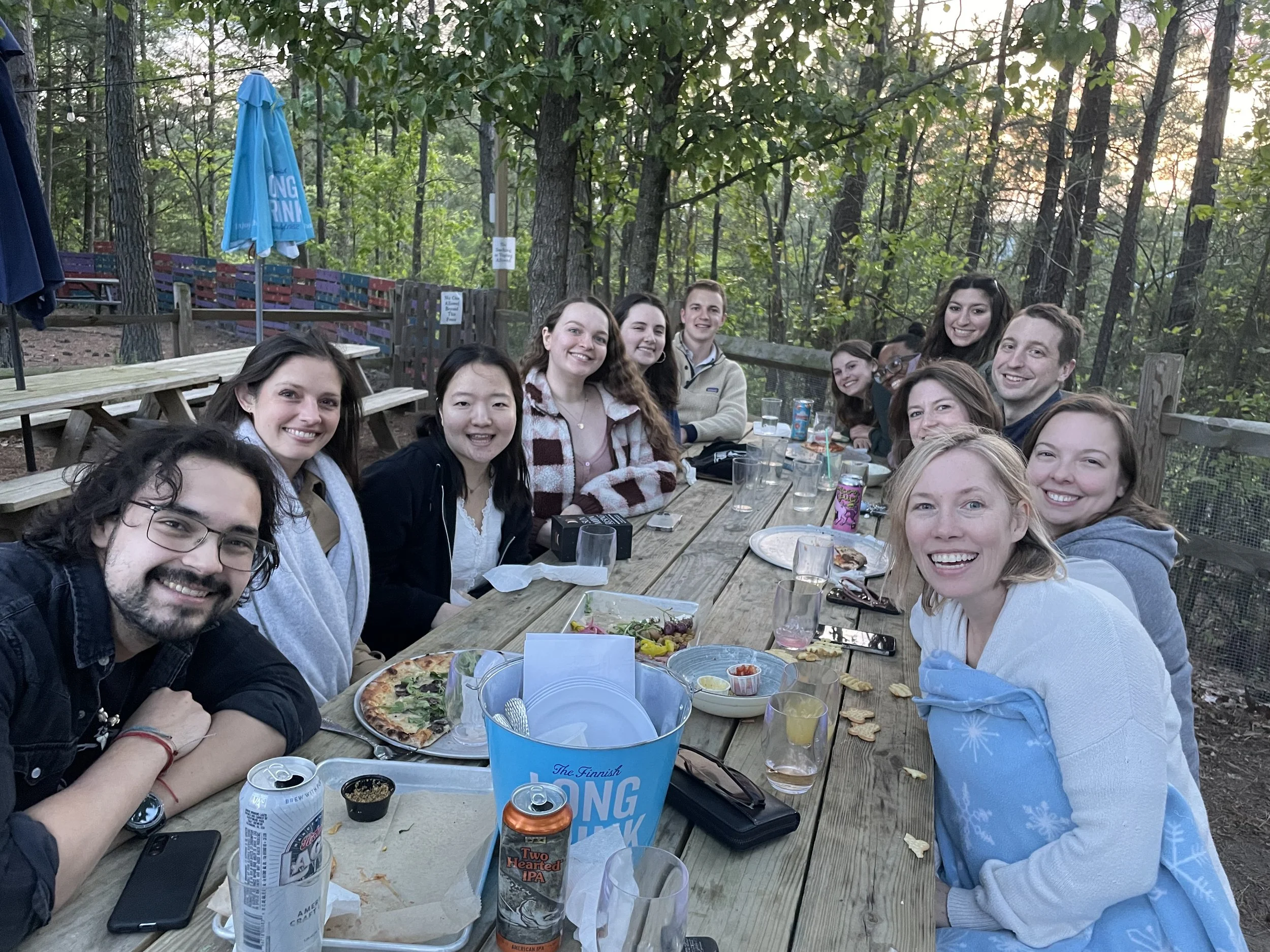 Group of people smiling and sitting at a picnic table in a wooded outdoor area during daylight, enjoying pizza and drinks.