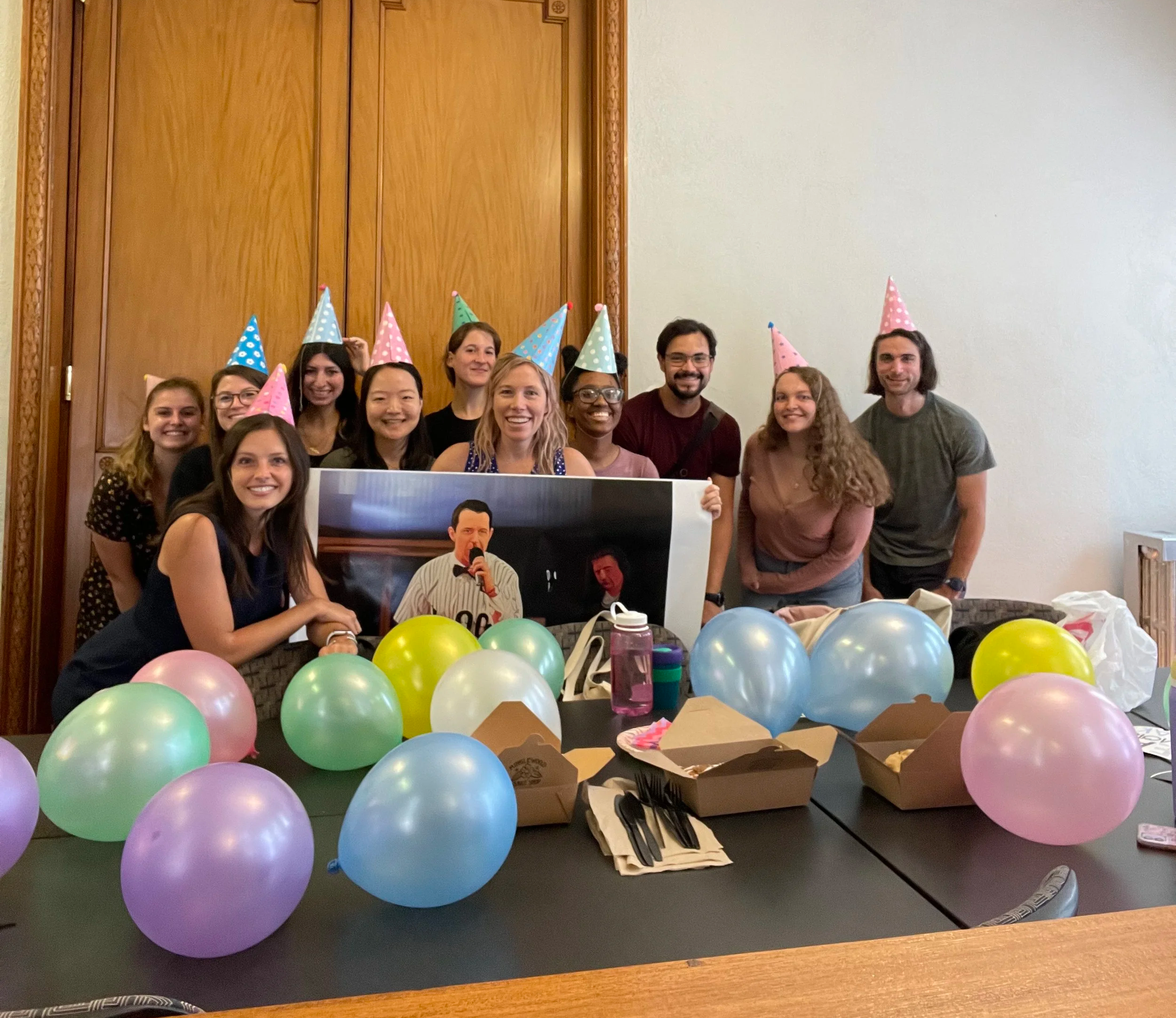 Group of people celebrating, wearing colorful party hats, surrounded by balloons, with a table filled with party food and decorations, in a room with wood-paneled door and chandelier.