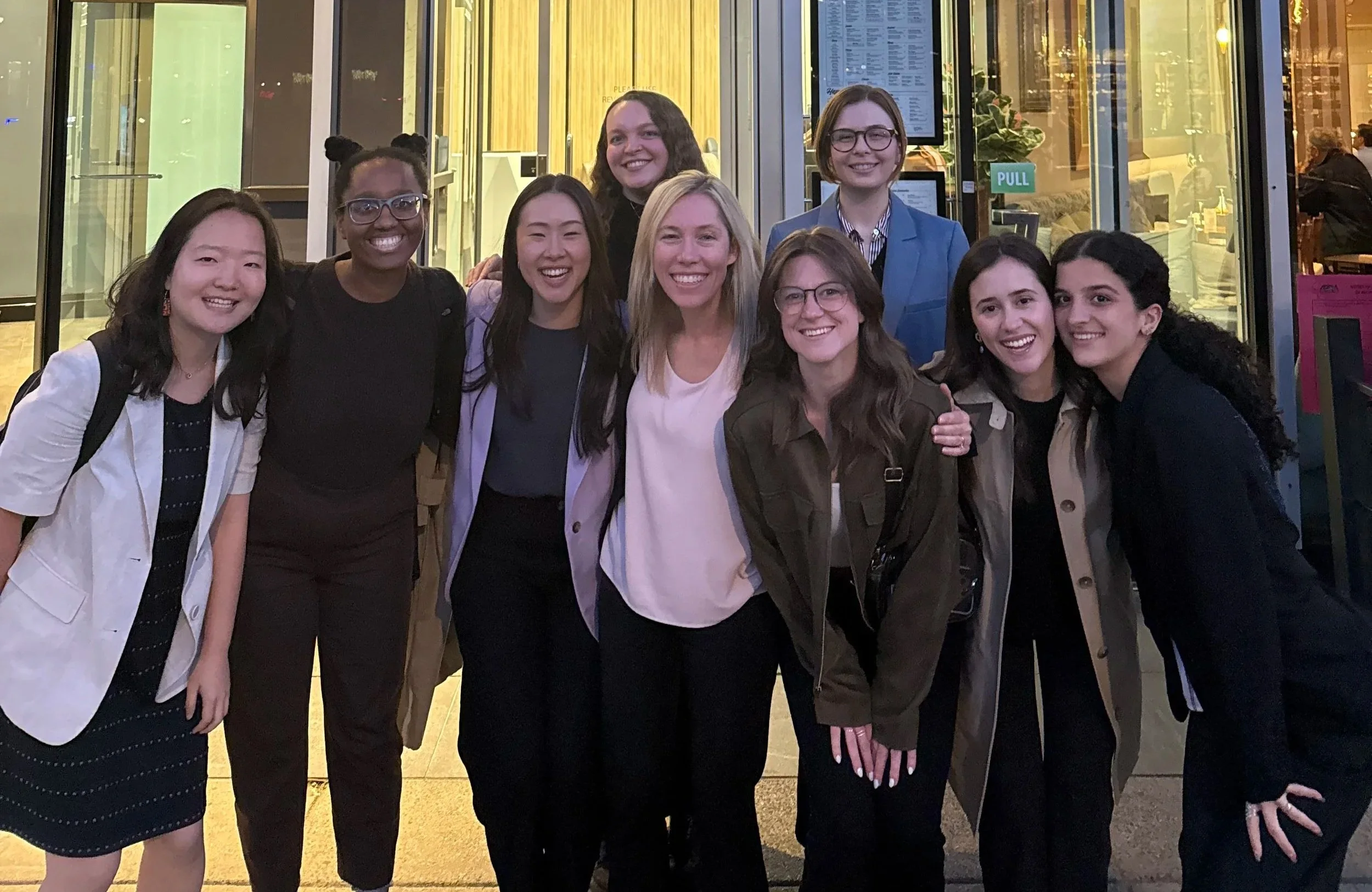 Group of ten women smiling and posing together outside a building at night, some wearing glasses and casual or business casual attire.